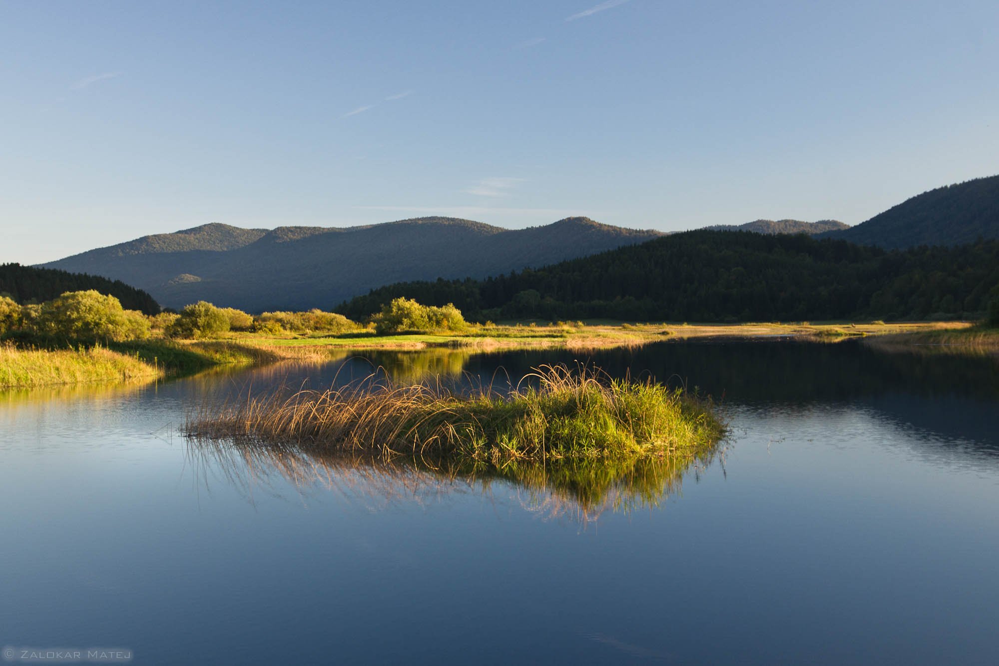 A serene river landscape with calm water reflecting the surrounding greenery, hills, and mountains under a clear blue sky.