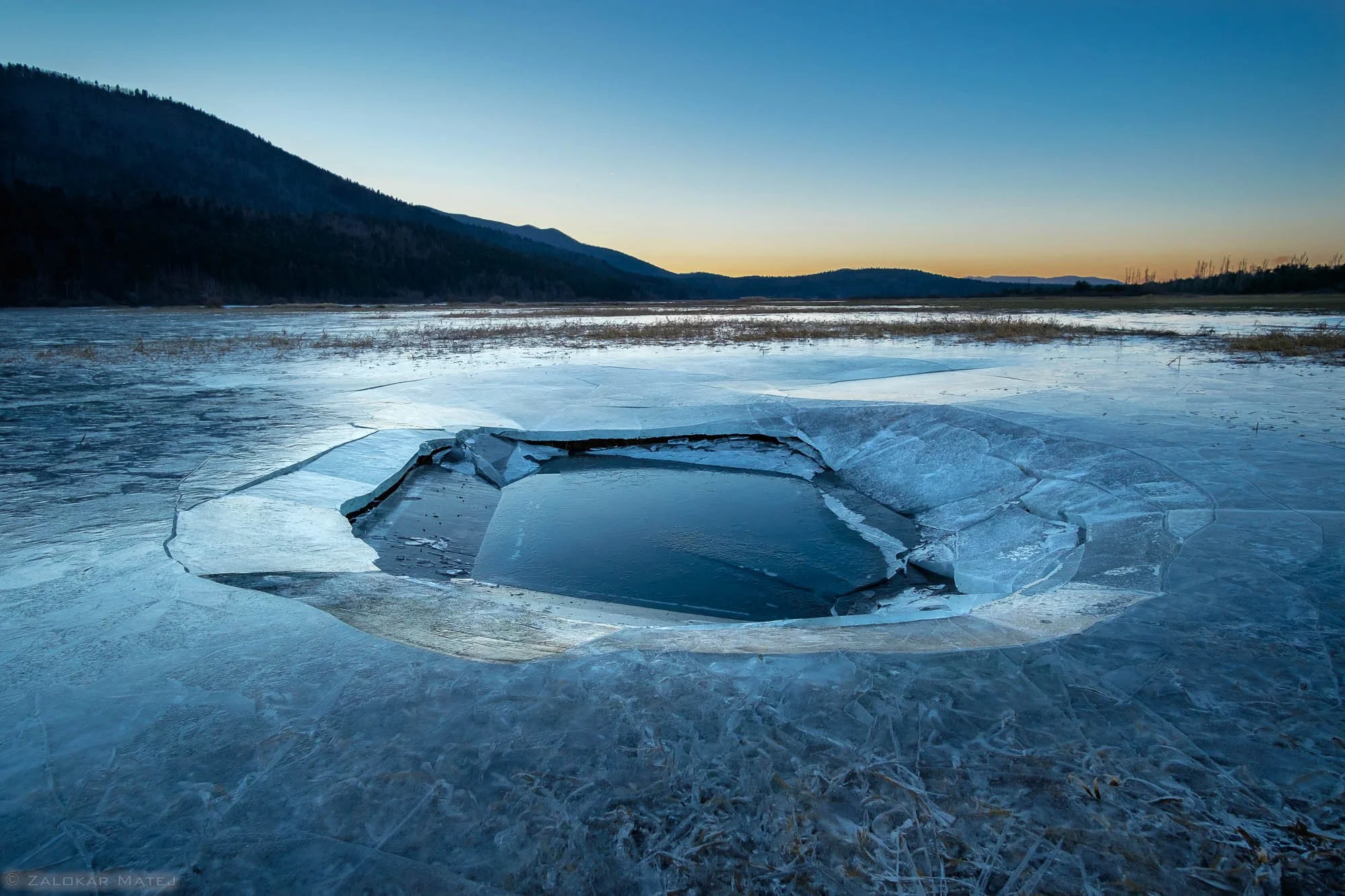 A frozen lake with a large hole in the ice revealing open water, surrounded by icy surface and distant hills under a sunset sky.