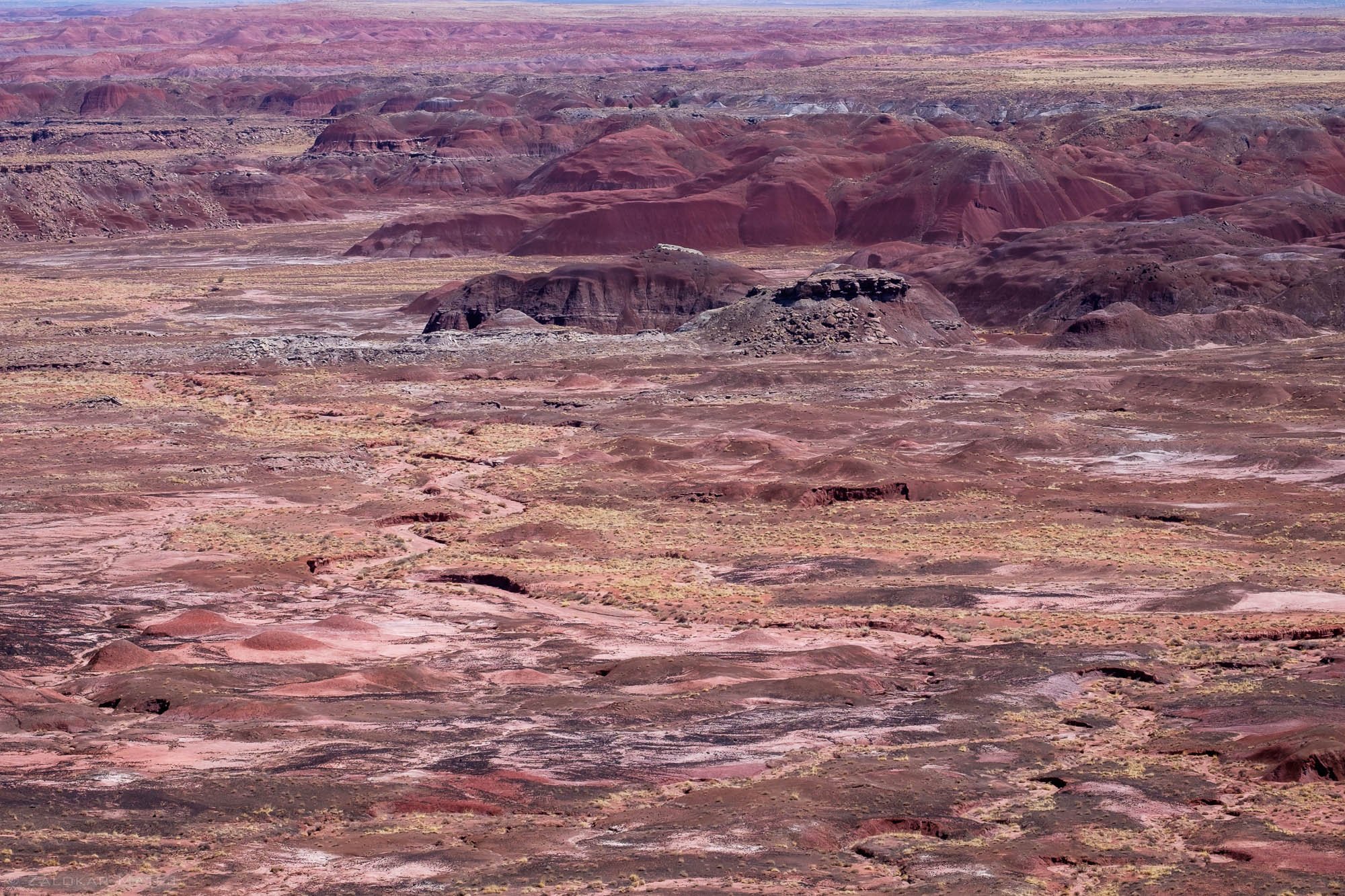 A vast desert landscape with layered, reddish-brown rock formations and mesas under a hazy sky.