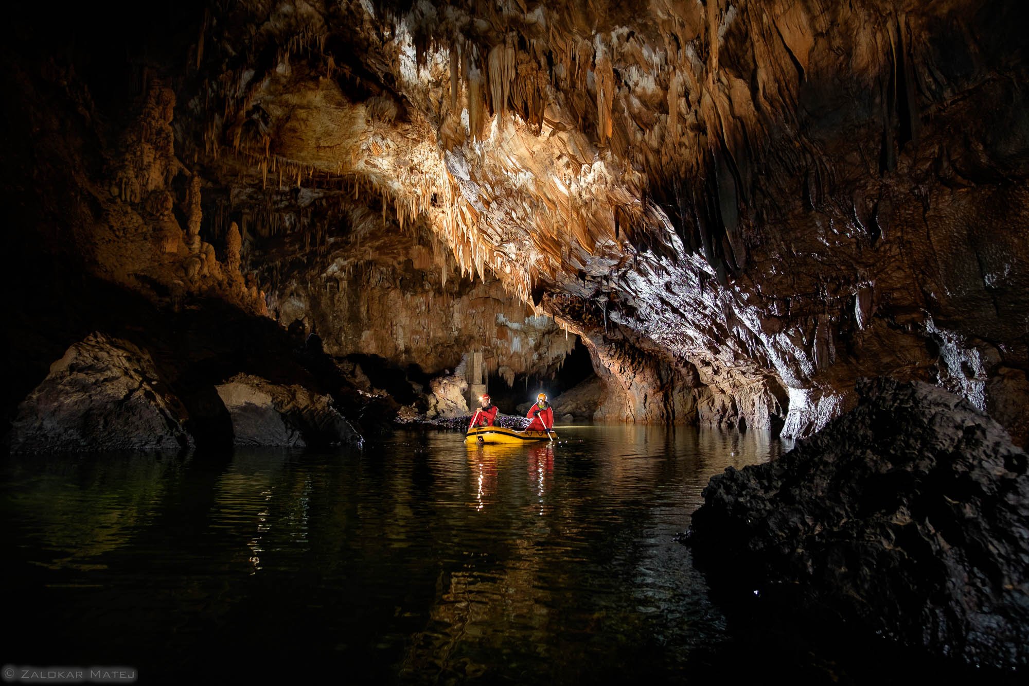 Two people in red life jackets on a yellow kayak exploring a dark cave with stalactites hanging from the ceiling, illuminated by artificial lighting.