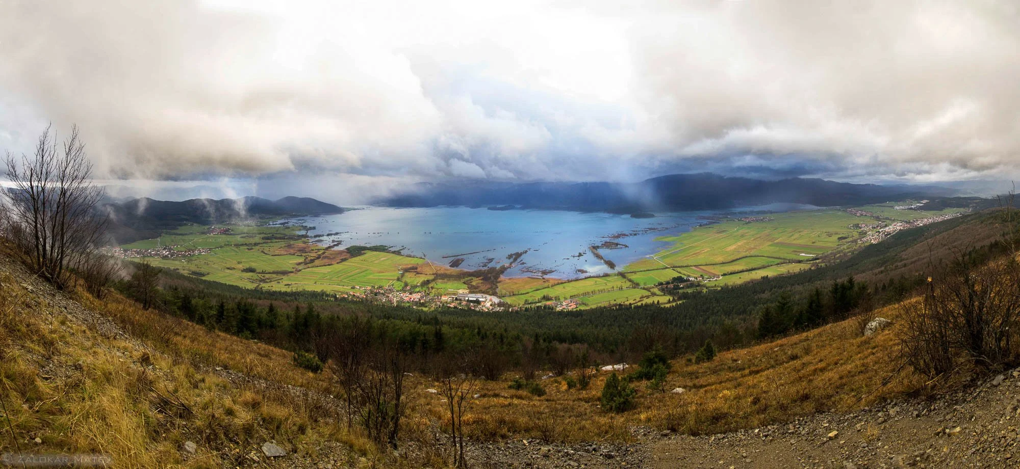 A panoramic view of a lake surrounded by green fields, small villages, and distant mountains under a cloudy sky.