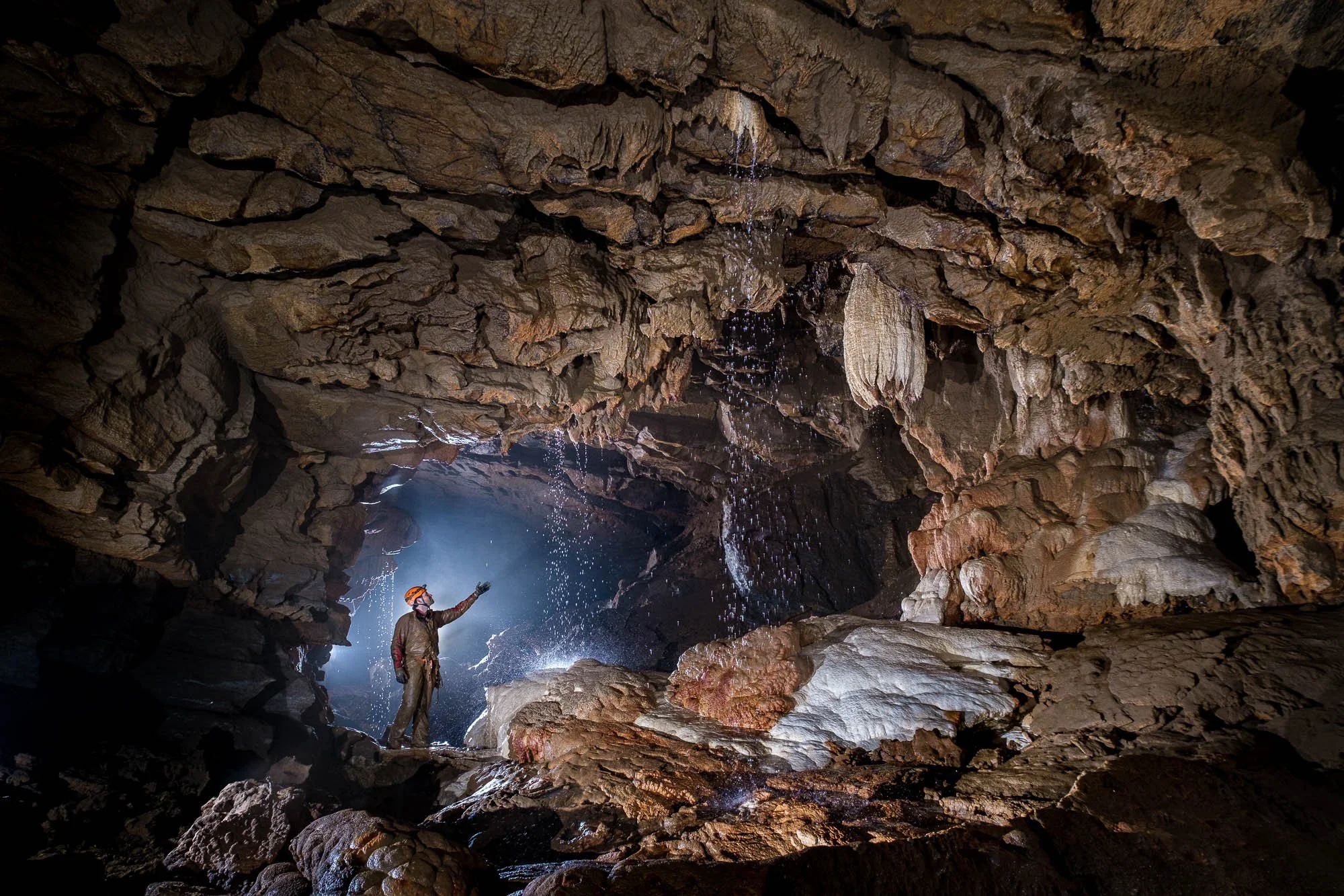 A caver wearing a helmet with a headlamp standing inside a dark cave with stalactites and stalagmites, and a waterfall of water droplets falling from the cave ceiling.