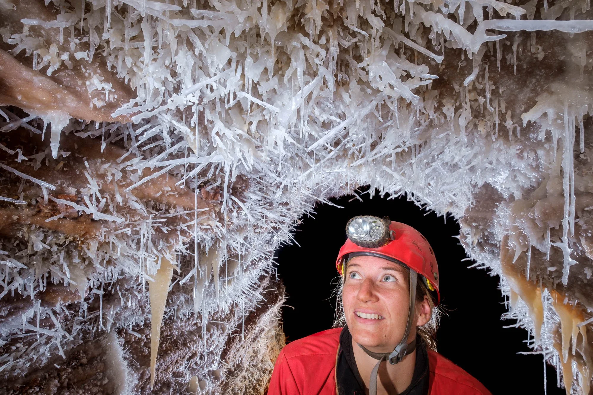A woman wearing a red helmet with a headlamp exploring a cave with ice formations.