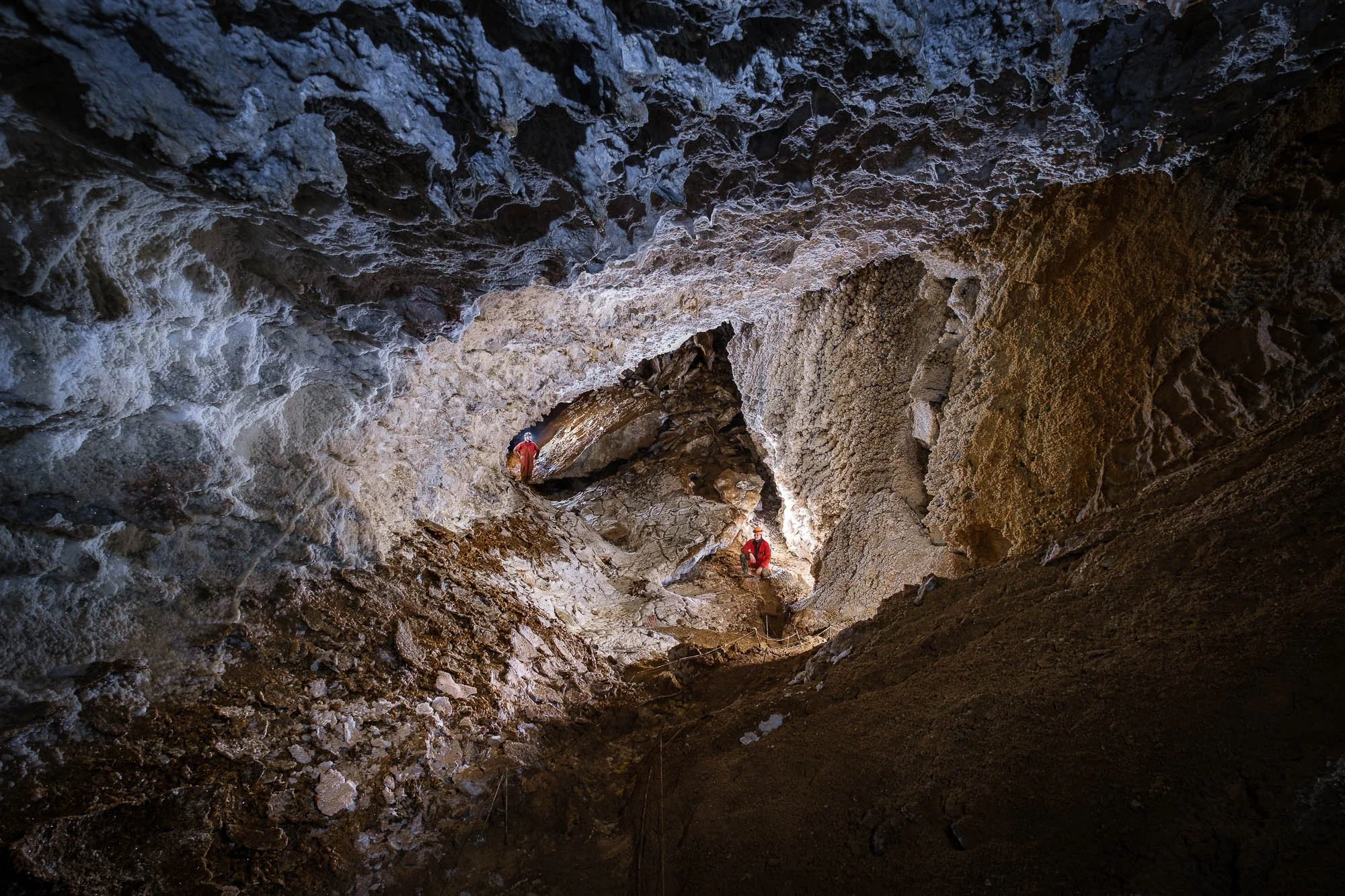 Two miners in red suits and helmets working inside a dark, rocky cave with uneven walls and formations.