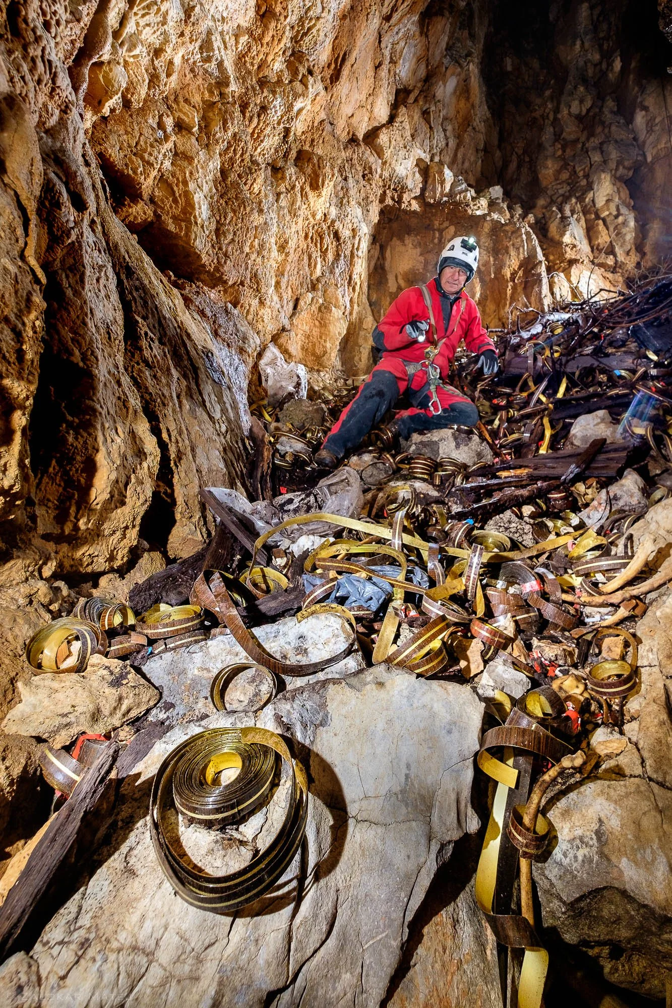 A person in safety gear and helmet sitting among a large pile of mining conveyor belts inside a rocky cave.