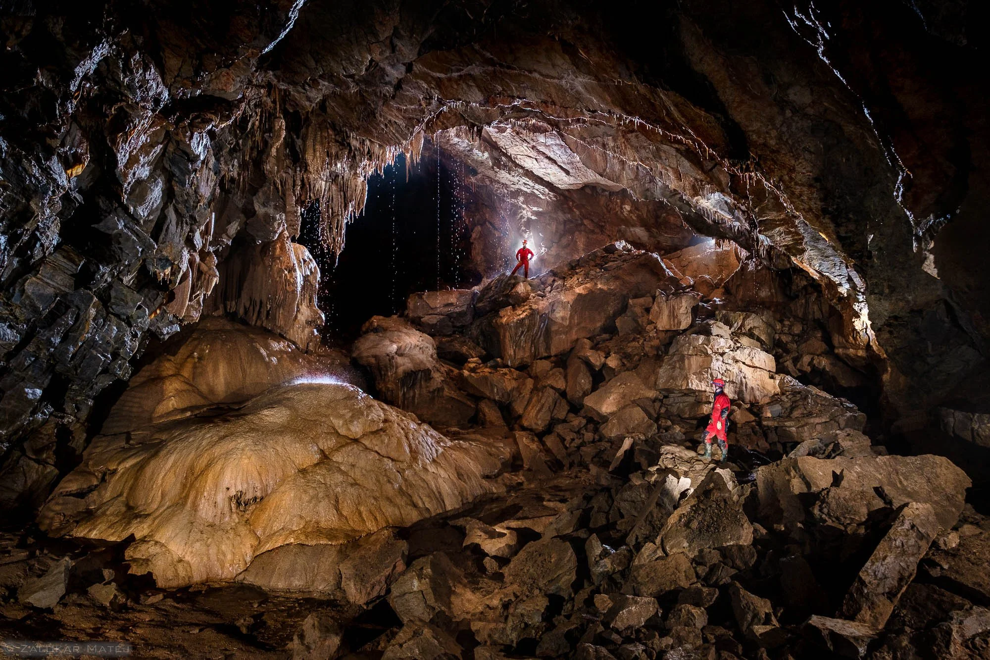 Two explorers in a large, dark cave with stalactites and uneven rocky terrain, one standing on rocks in the background illuminated by a light, the other on rocks in the foreground.
