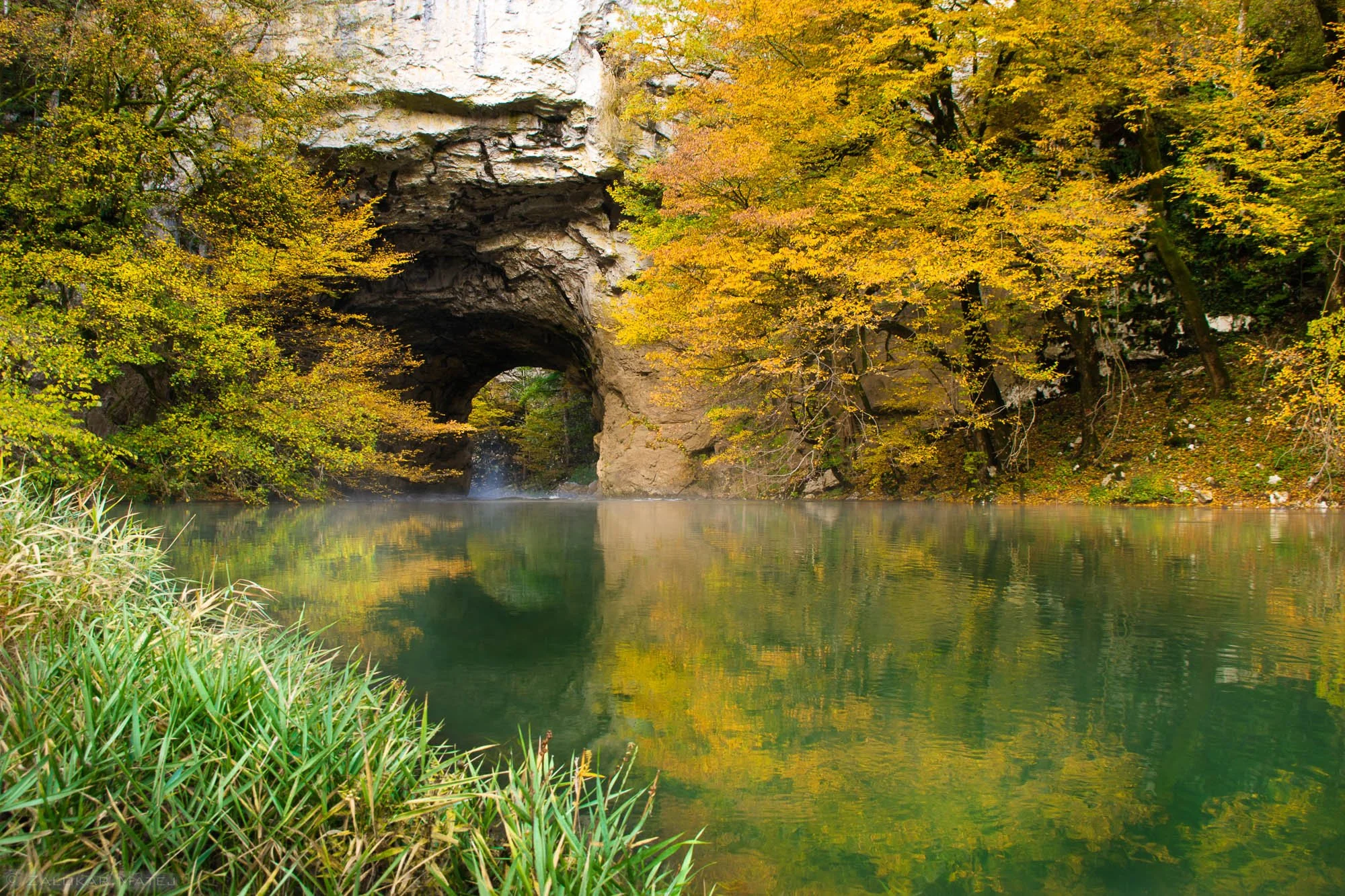 A scenic view of a river with calm green water, surrounded by autumn-colored trees with yellow and orange leaves, and a large rock formation with a natural archway in the background.