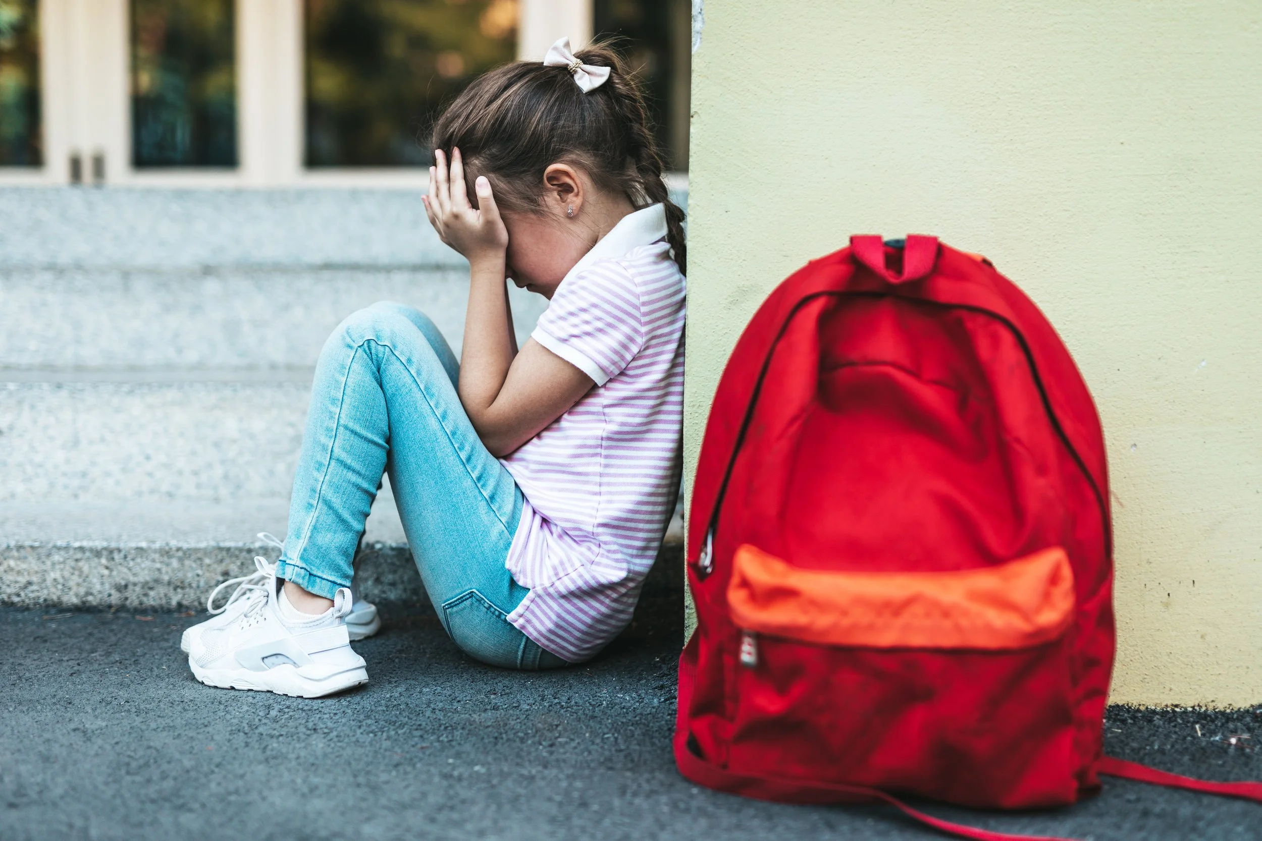 A young girl sitting on the ground with her head in her hands, appearing upset, next to a red backpack outside a building.