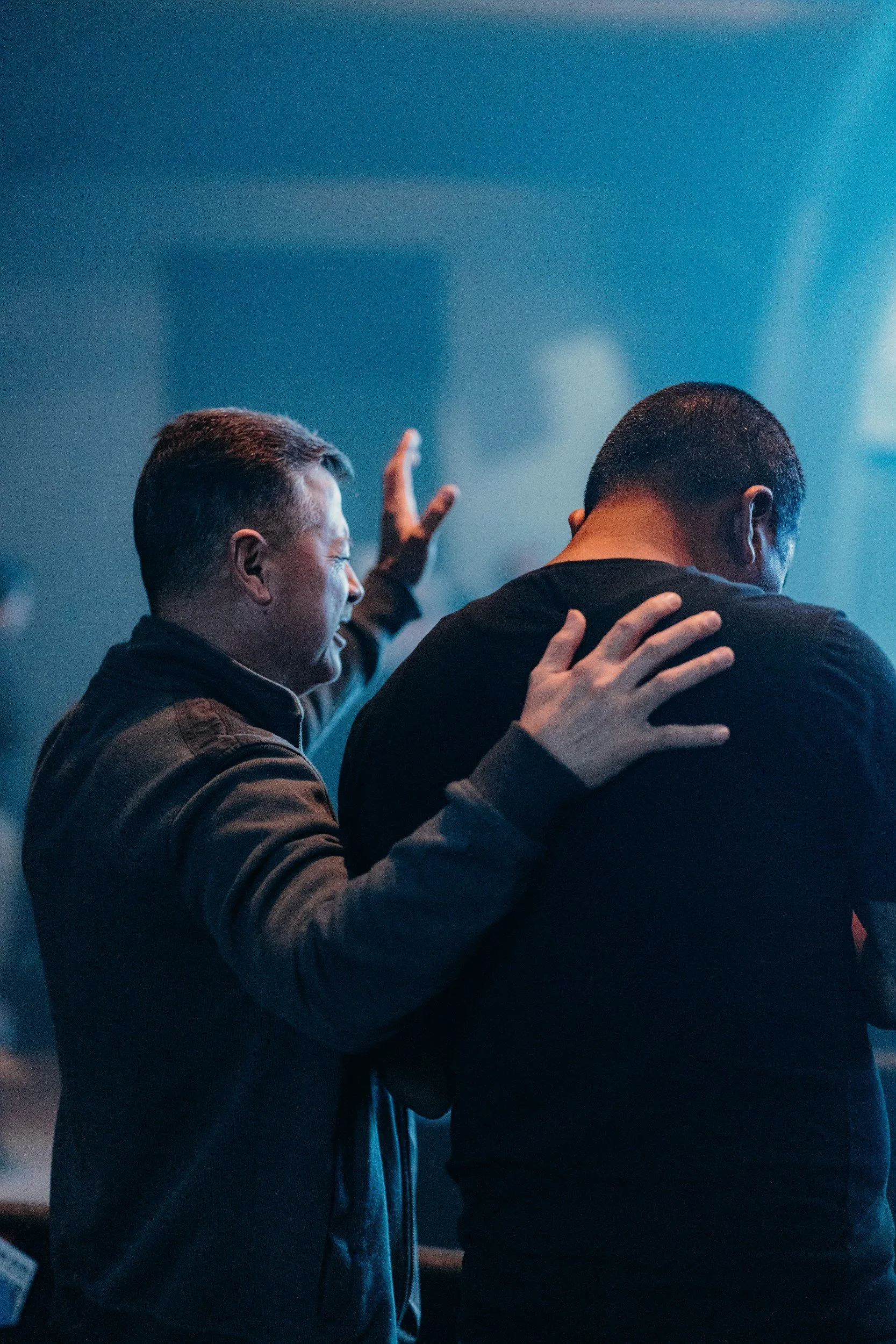 A man with closed eyes and a joyful expression prays with his hand raised, while another man offers comfort with a hand on his shoulder in a dimly lit setting with blue lighting.