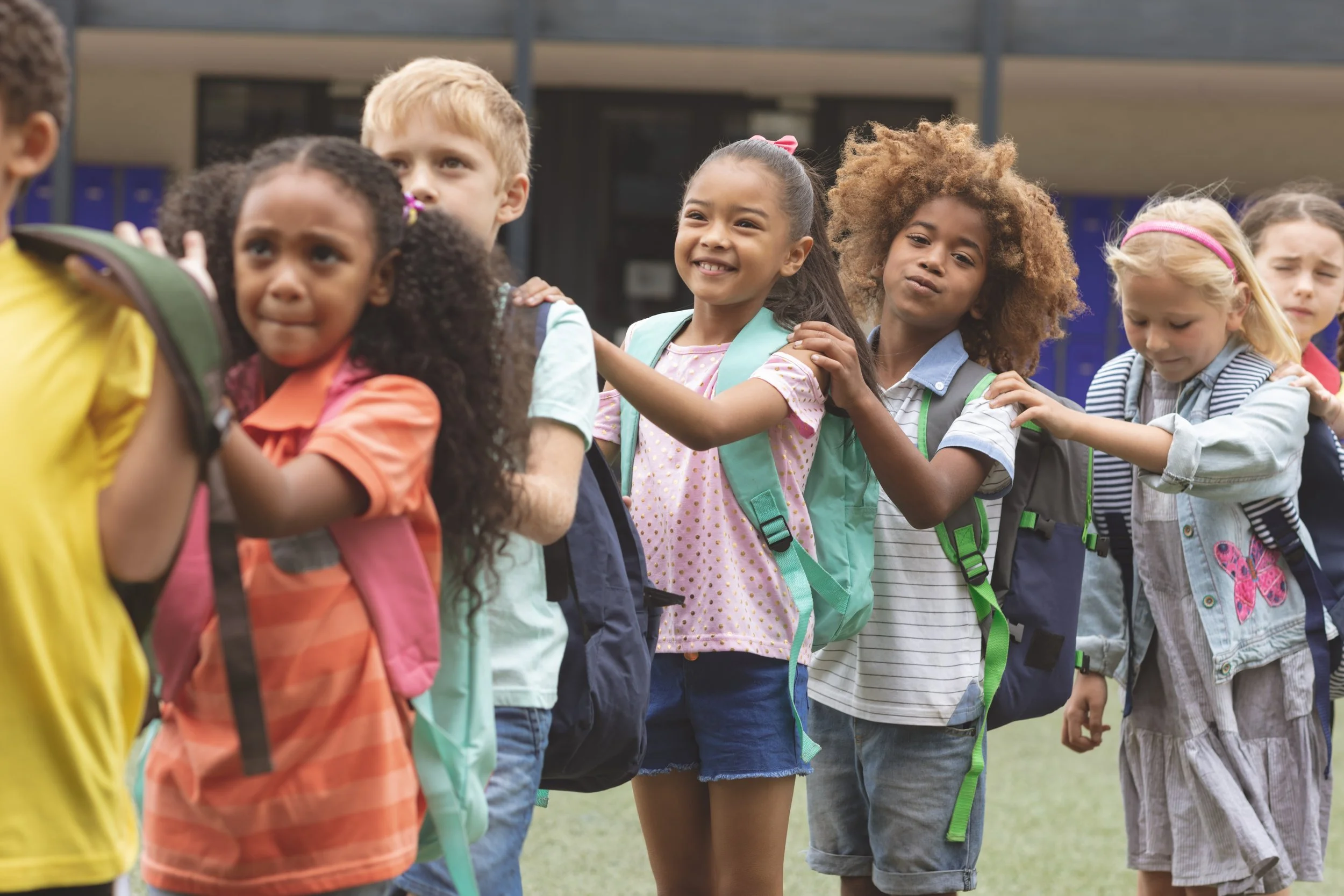 A line of diverse children standing outside, holding onto the shoulders of the child in front of them, at a school campus with lockers in the background.