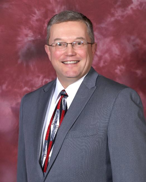A smiling man in a gray suit, white shirt, and patterned tie, standing against a maroon textured background.