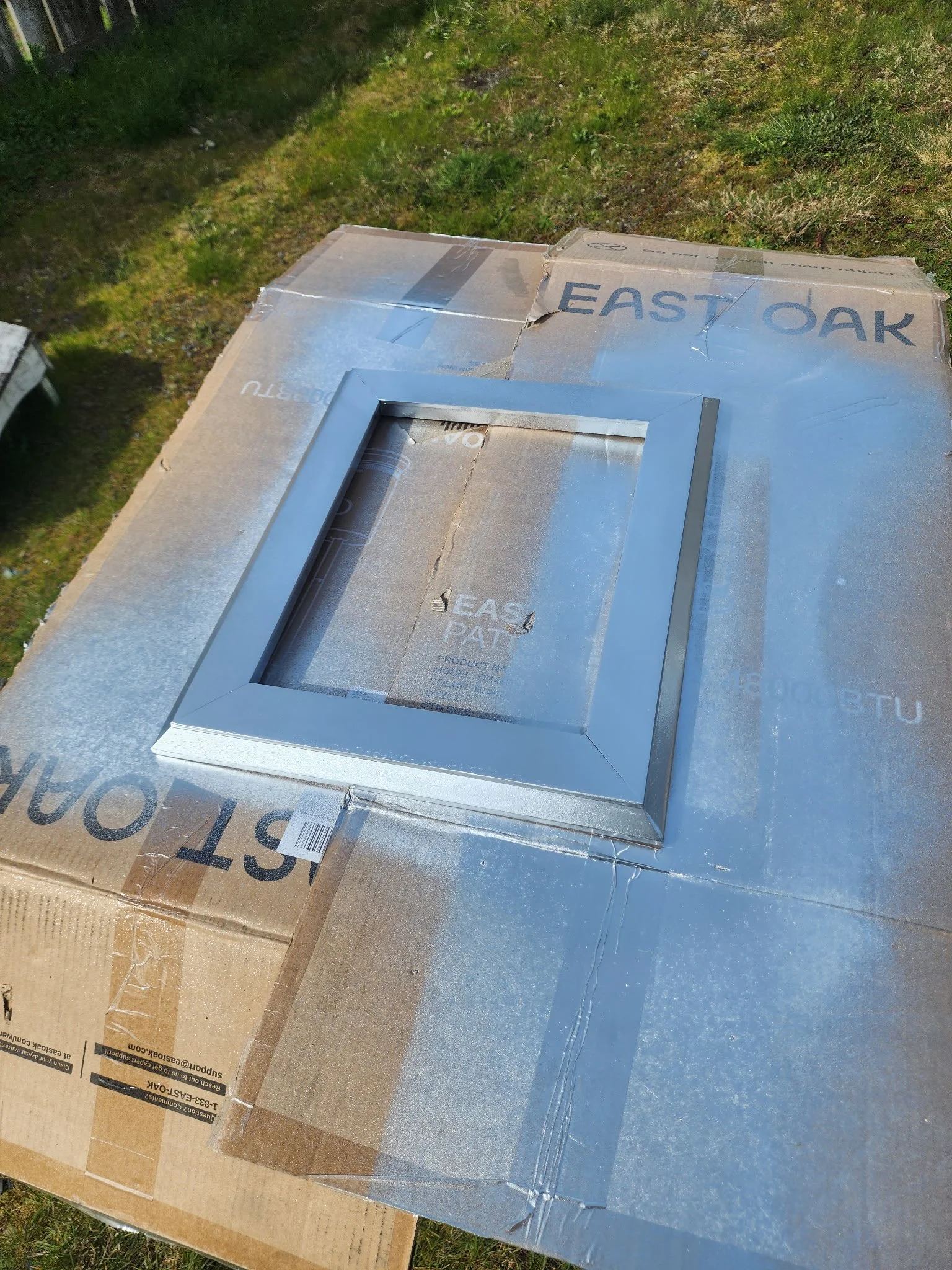 A cardboard box with a metal frame on top, placed on a larger cardboard surface outdoors with grass and ground visible in the background.