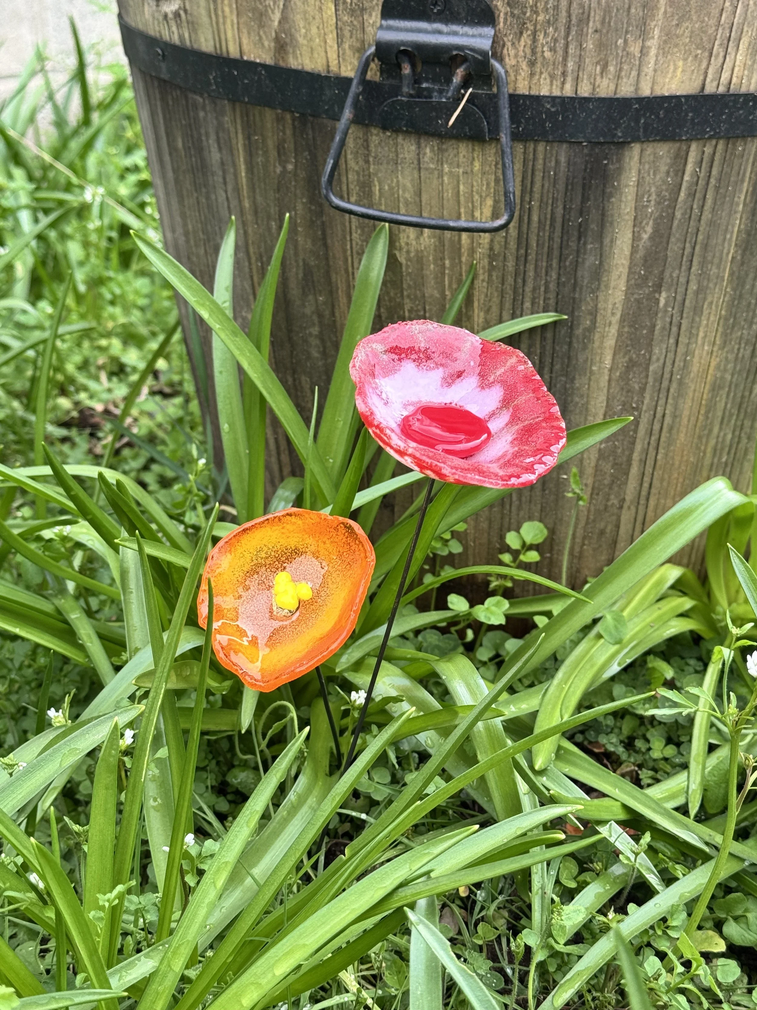 Colorful garden flowers with two artificial flowers, one red and white, and one orange and yellow, in front of a wooden post with a metal latch.