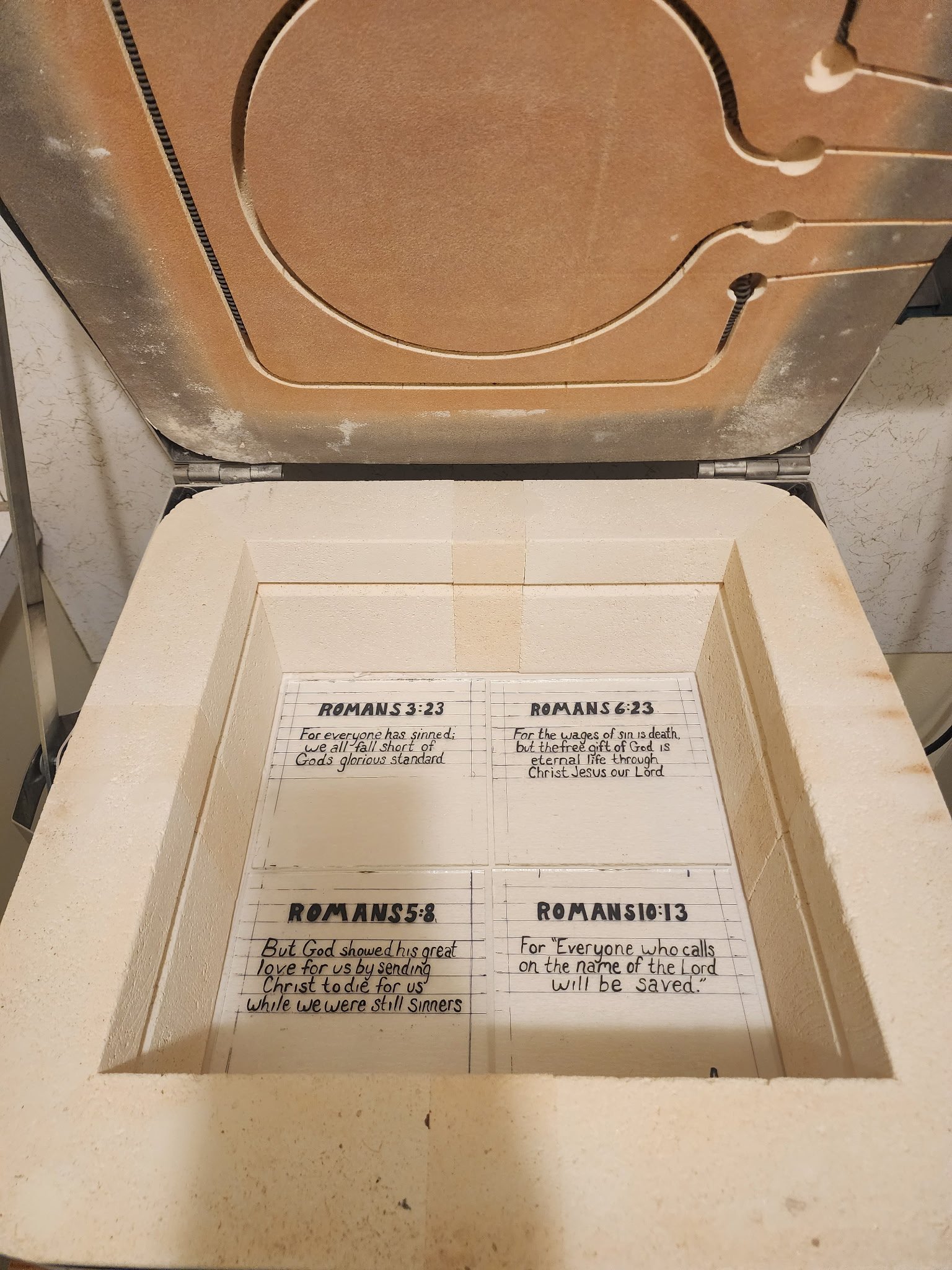 Open white box with handwritten notes containing Bible verses, with the lid open, in a room with a textured wall in the background.