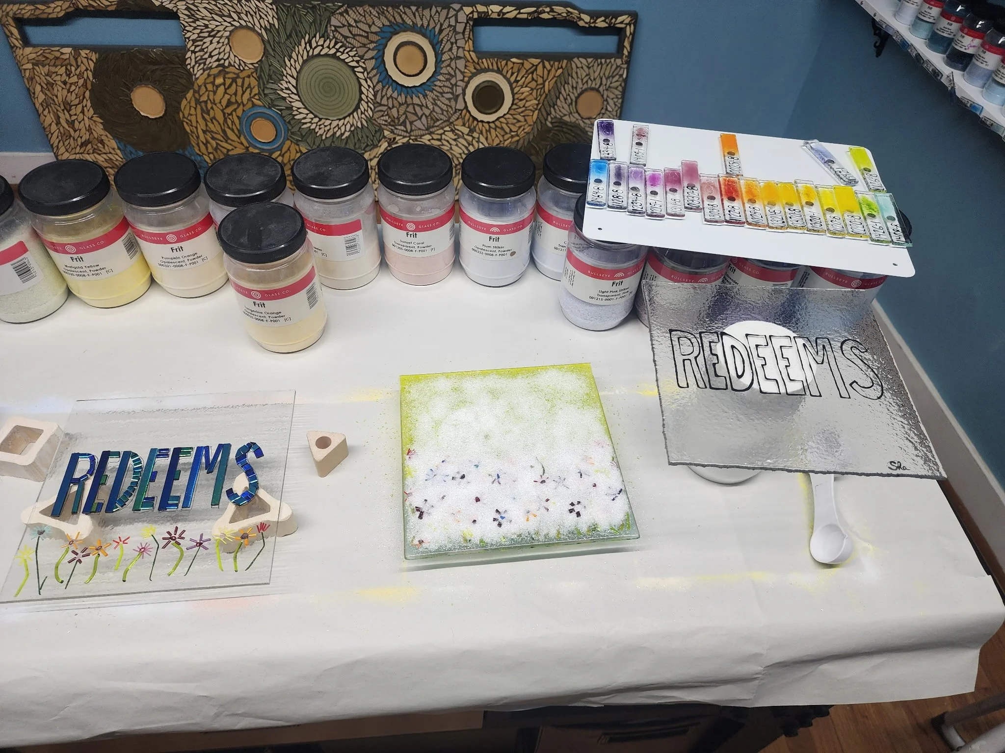 A display table with jars of paint, a color palette, and handmade signs that say 'REDEEMS.' The jars are labeled with colors and are arranged across the table, with a color chart on a raised platform. There are various art supplies and decorative ite