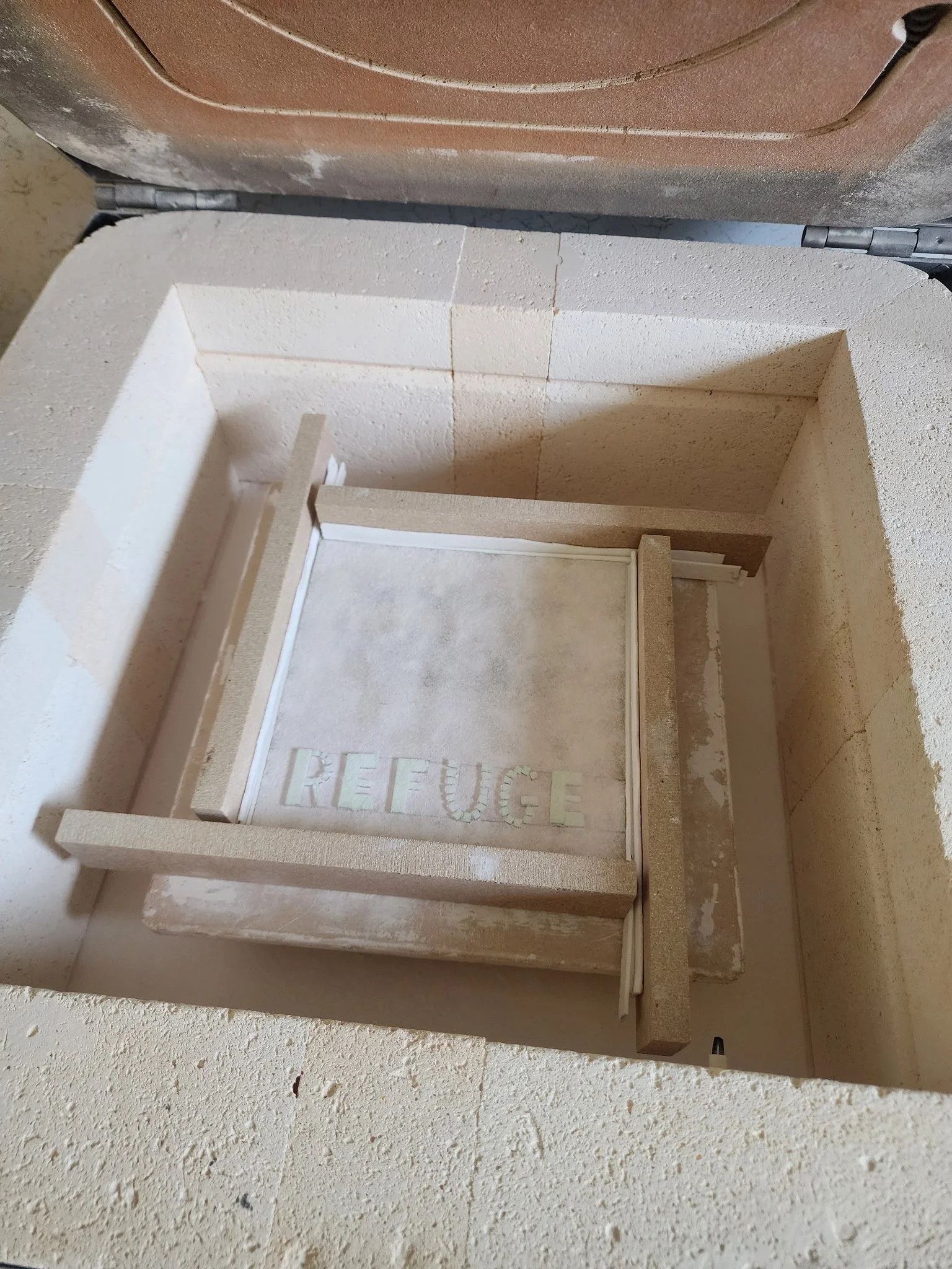 A small, beige-colored, enclosed space with the word 'REFUGE' written on its floor, surrounded by stone or concrete walls, with a brown lid or cover at the top.