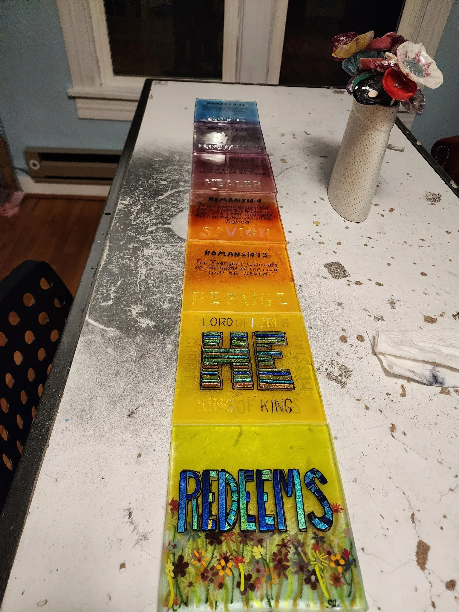 A row of colorful glass tiles with biblical quotes and phrases arranged on a white, painted wood table next to a floral-patterned vase with artificial flowers.