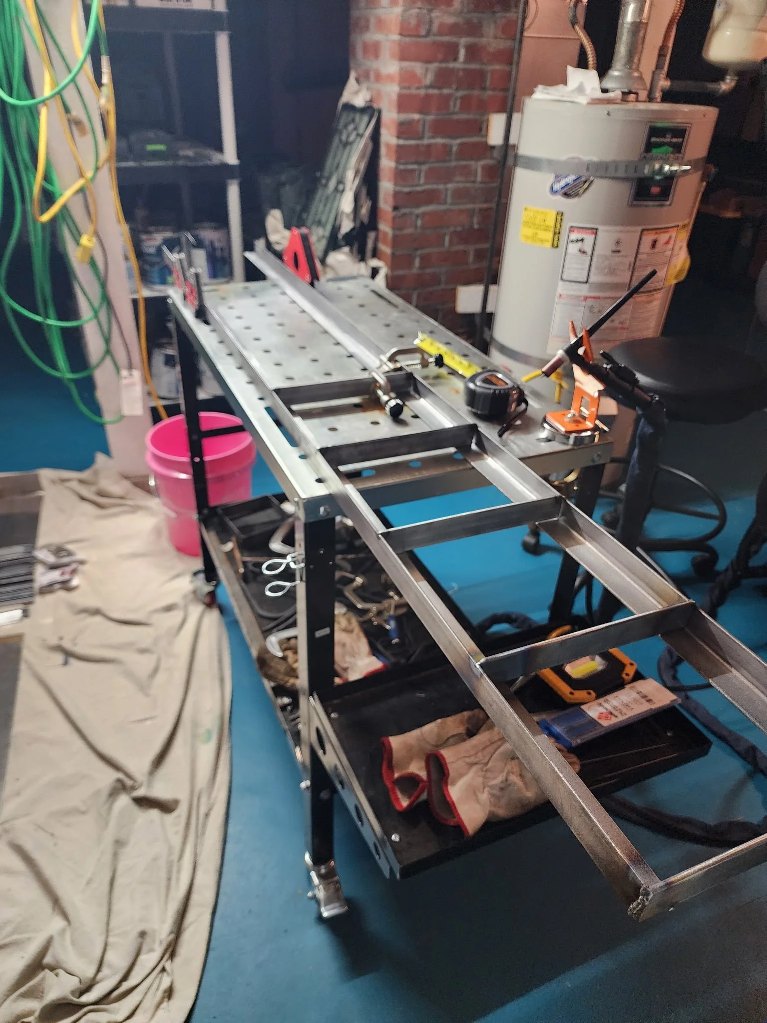 A workbench with tools and equipment, including a tape measure, a drill, and a metal frame, in a workshop setting. There are gloves on the lower shelf, a pink bucket nearby, and a water heater behind.