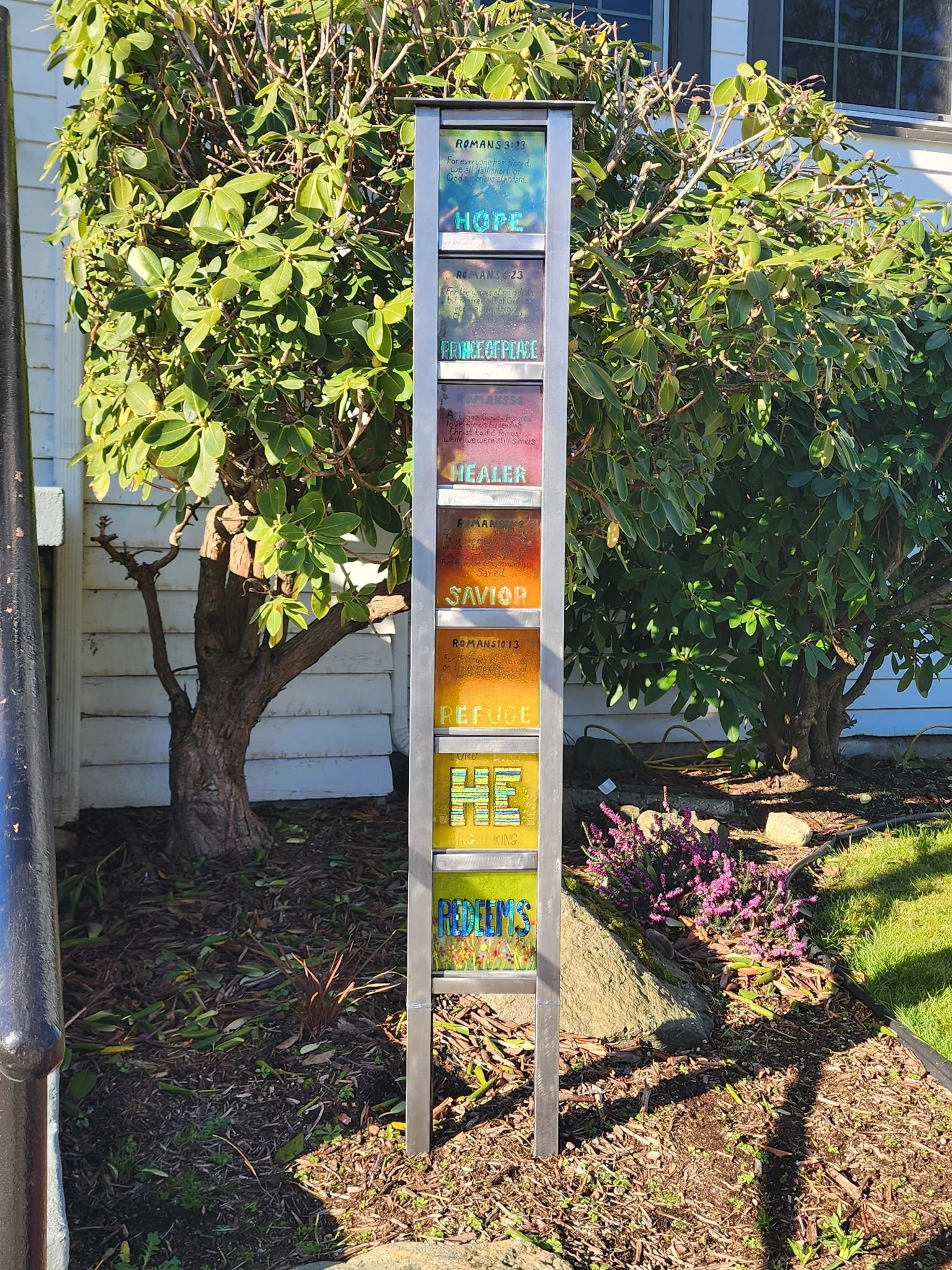 A tall, narrow sign with colorful, illuminated words situated outdoors in front of green shrubbery and flowers.