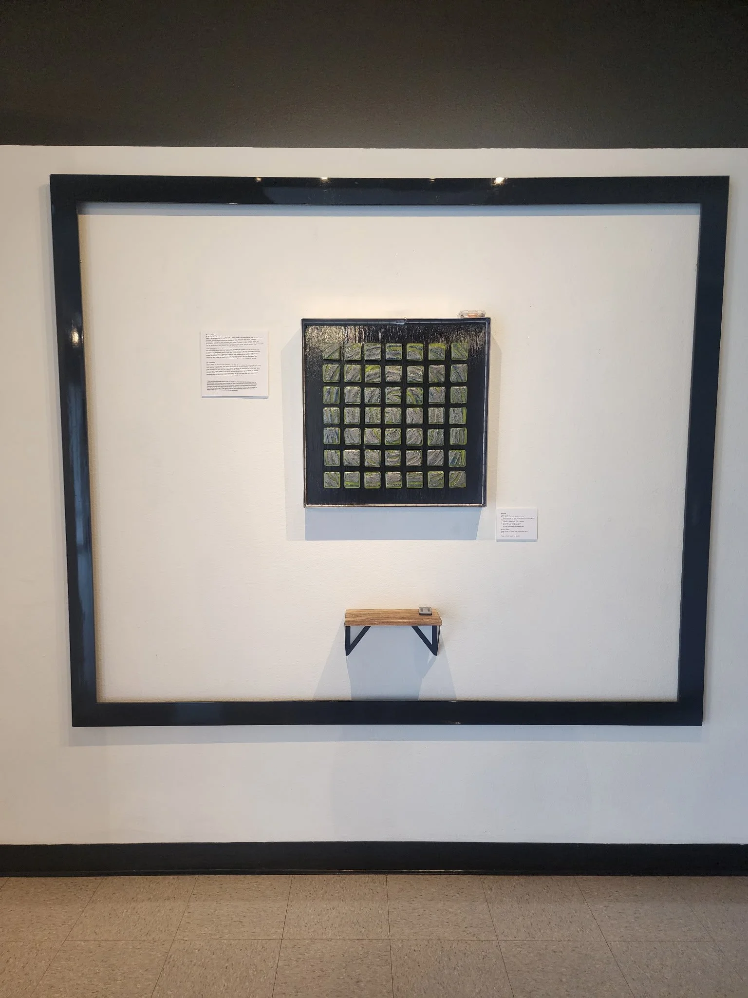 A framed artwork with square tiles painted with a landscape scene, displayed on a white wall with informational plaques, and a small wooden shelf below.