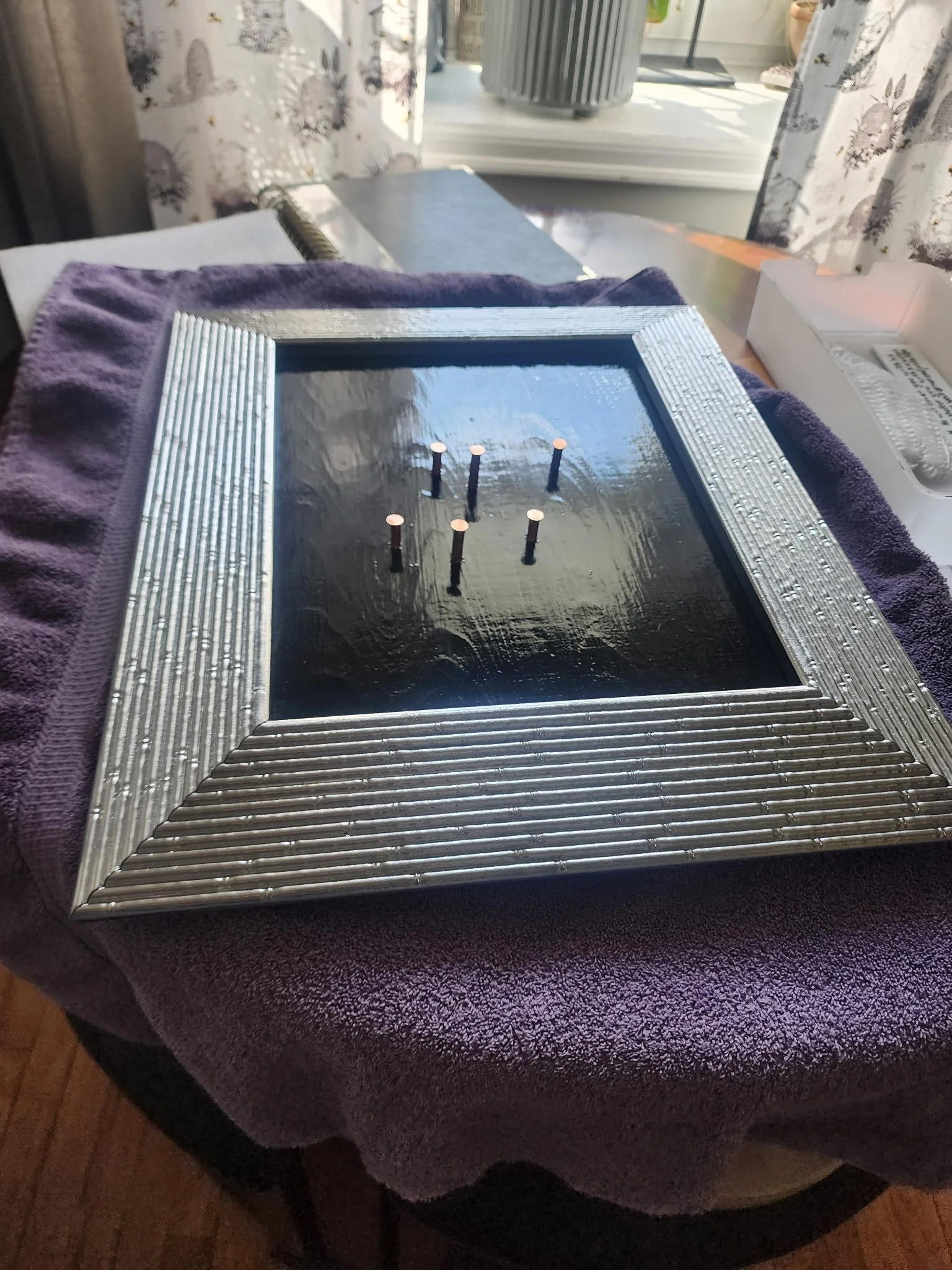 A square mirror with a silver, textured frame, placed on a purple towel on a table covered with a purple cloth. The mirror’s reflective surface shows five candles standing on a surface, with a window and curtains in the background.