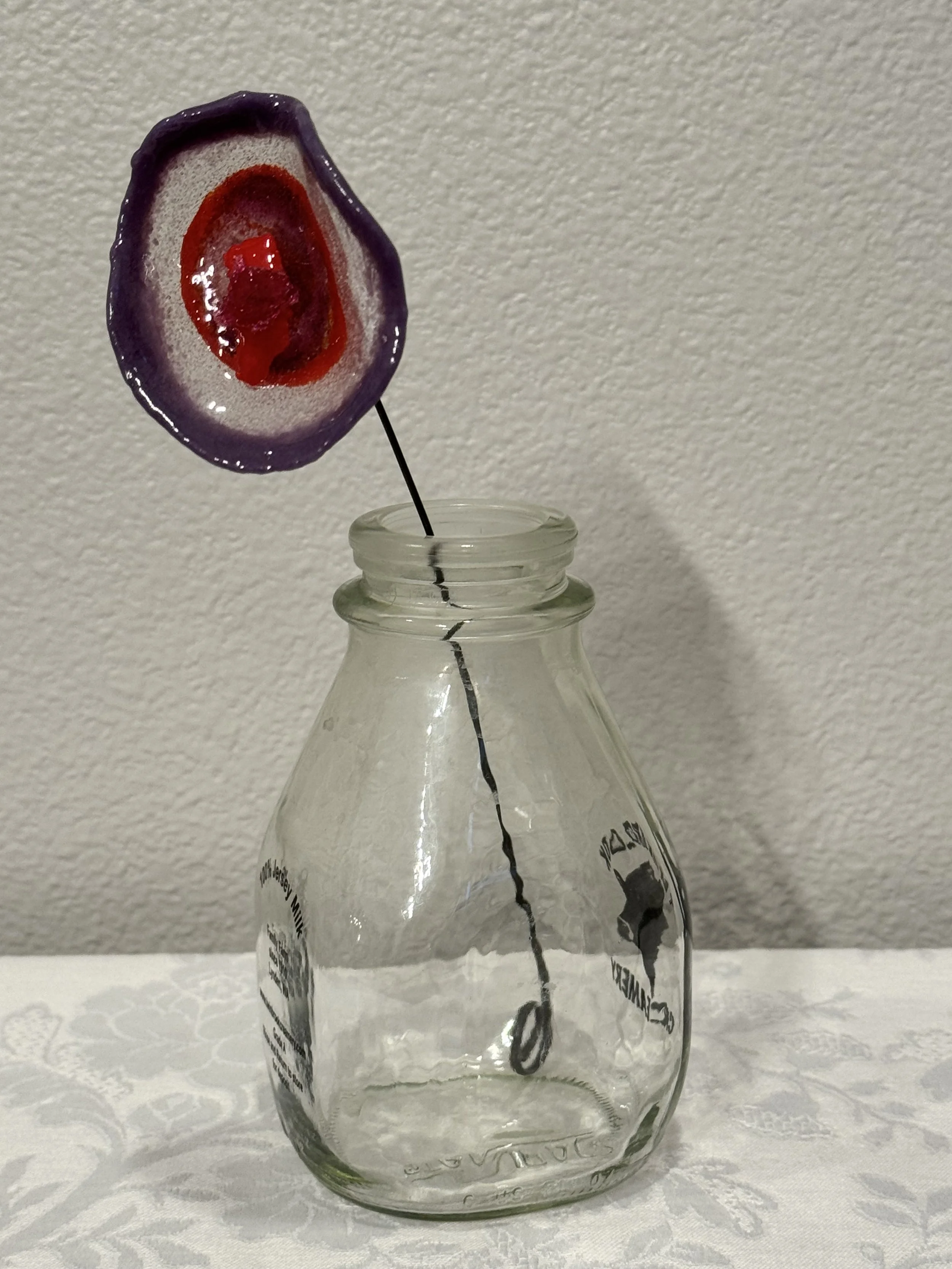 A clear glass Gatorade bottle with a black black straw inside it, decorated with a purple and red lollipop in front of a plain wall and a white tablecloth.