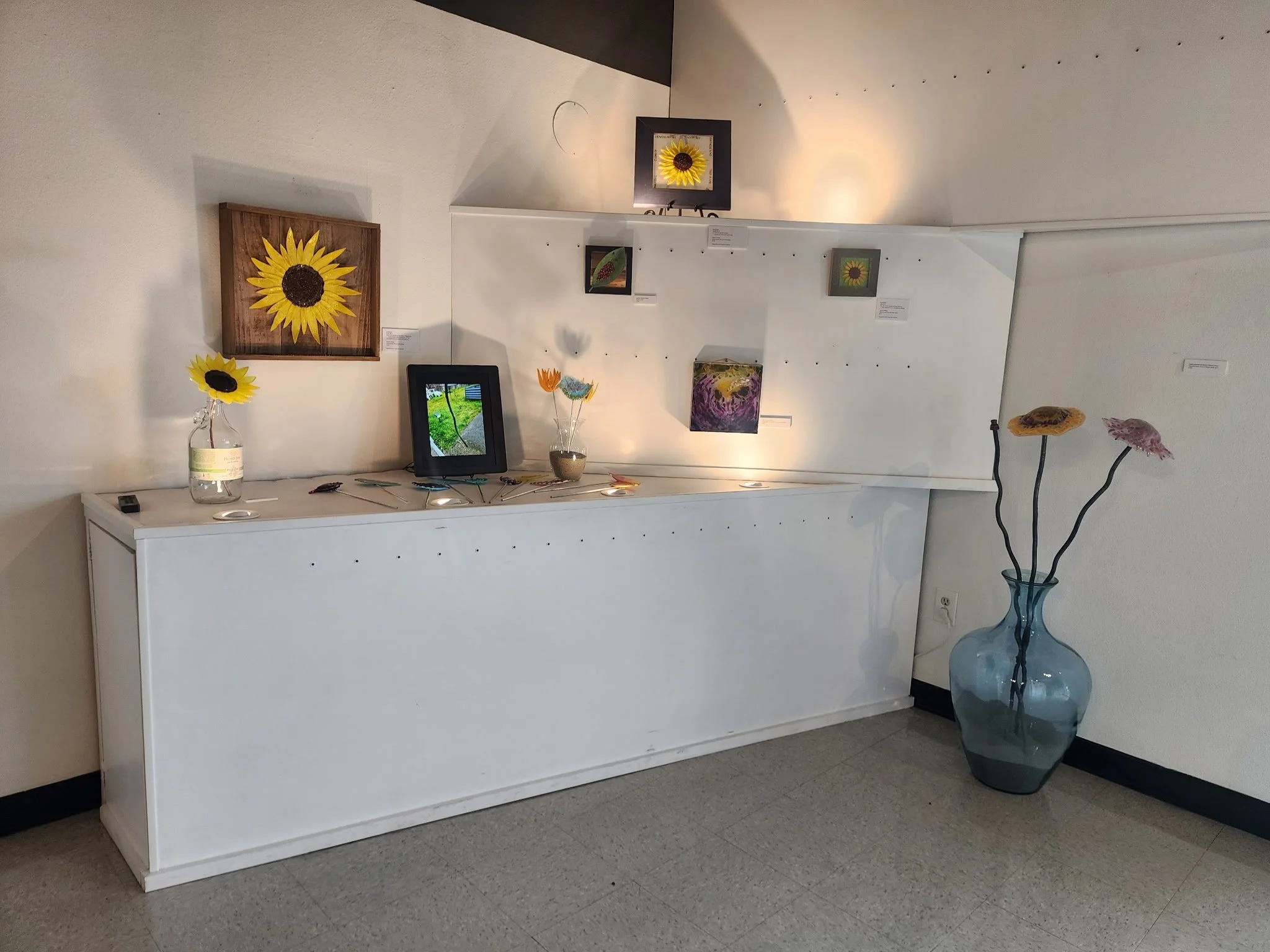 An art display with sunflower-themed artworks on the wall, a white cabinet with flower arrangements, and a large glass vase with three artificial flower stems.