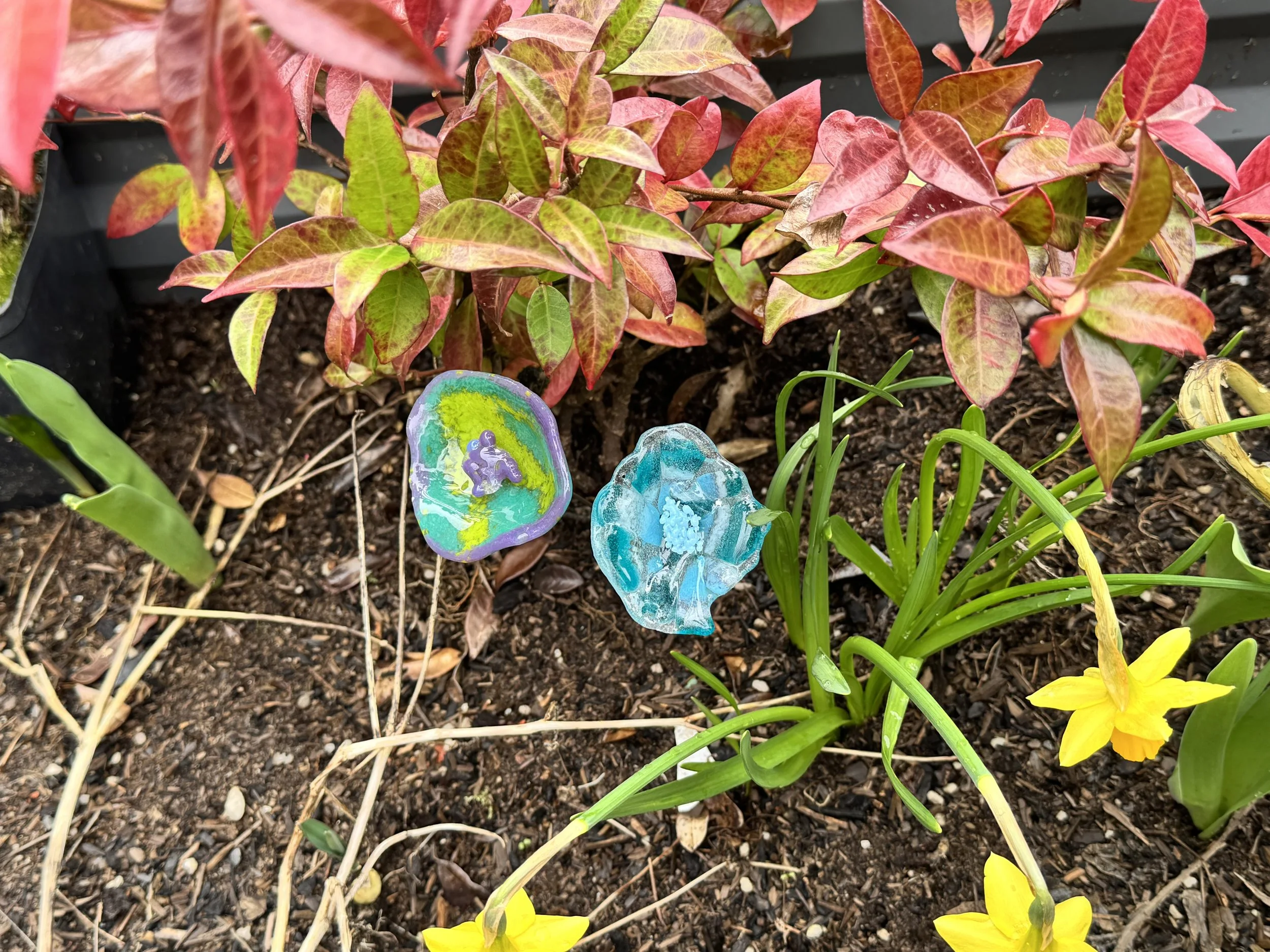 Colorful glass garden decorations among yellow daffodil flowers and red and green leafy plants in a garden bed.