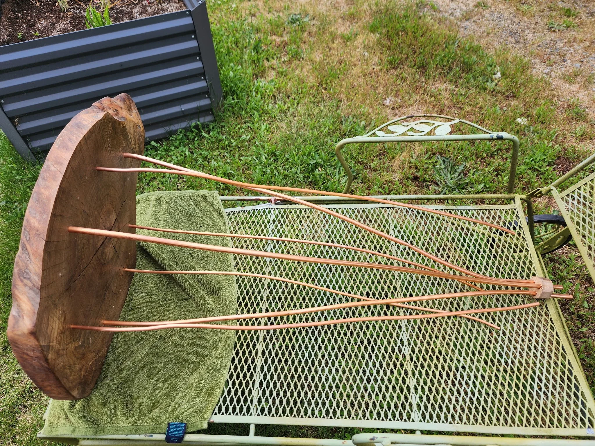Copper-colored garden stakes lying on a green metal outdoor table with a wood slice as a base and a green towel, outdoors on grass.