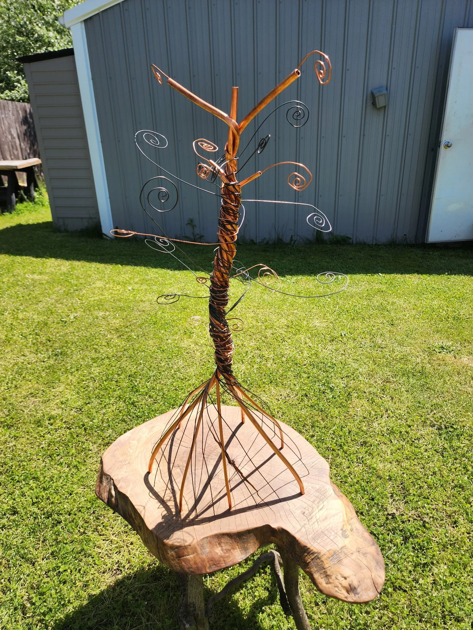 Wire sculpture of a tree with curled branches on a wooden pedestal in an outdoor yard.