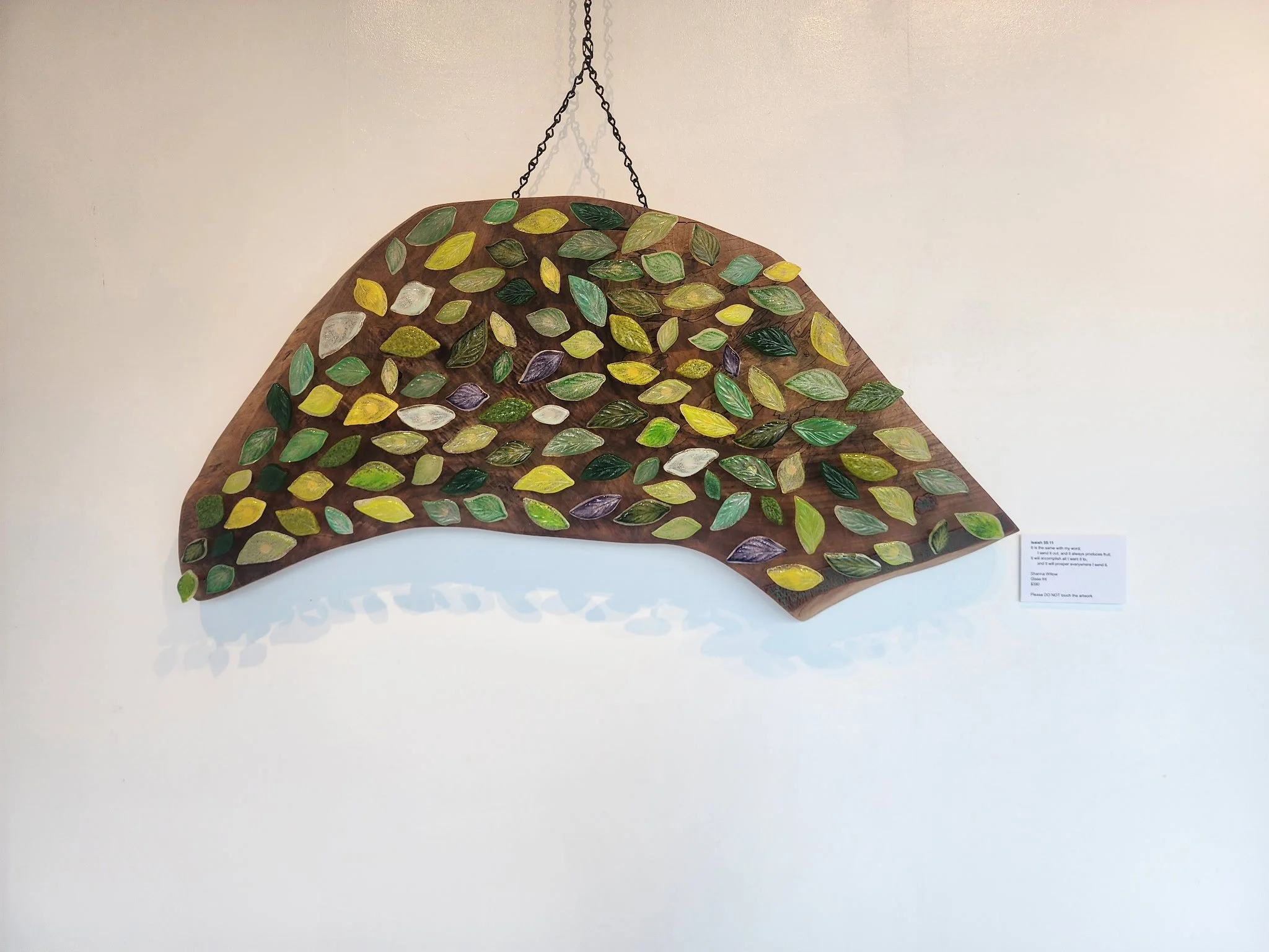 A hanging wooden art piece with painted green, yellow, and purple leaves on a white wall.