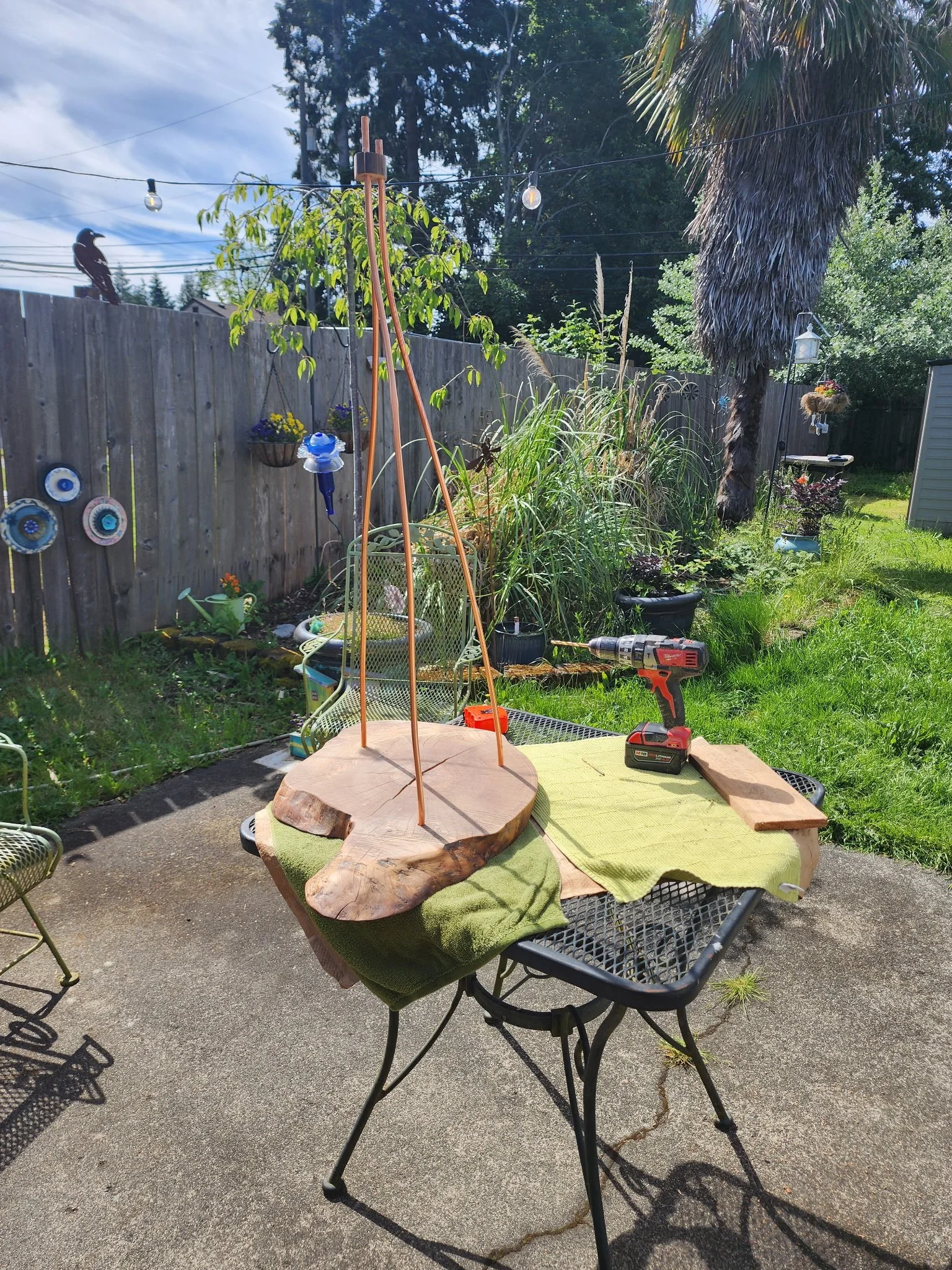 A backyard patio with a metal table, showing a woodworking project in progress. The project involves a round wooden base and four copper rods sticking out. There is a drill, a tape measure, and some fabric or towels on the table. The background inclu