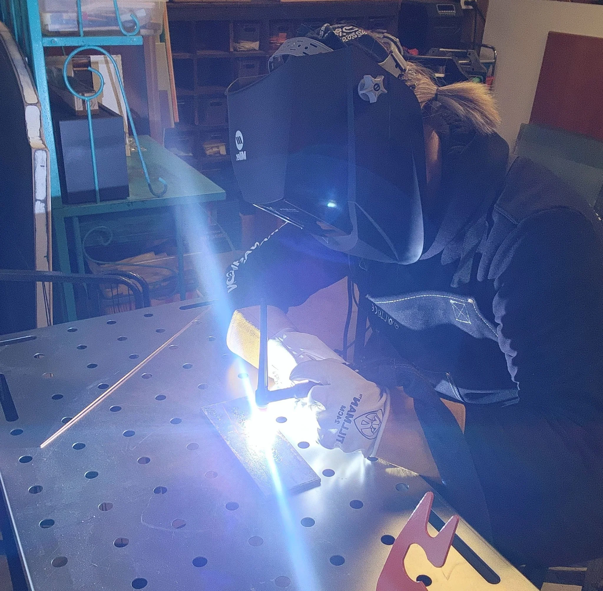 Person welding at a workbench with welding gear, wearing safety gloves and a welding helmet, emitting bright sparks.