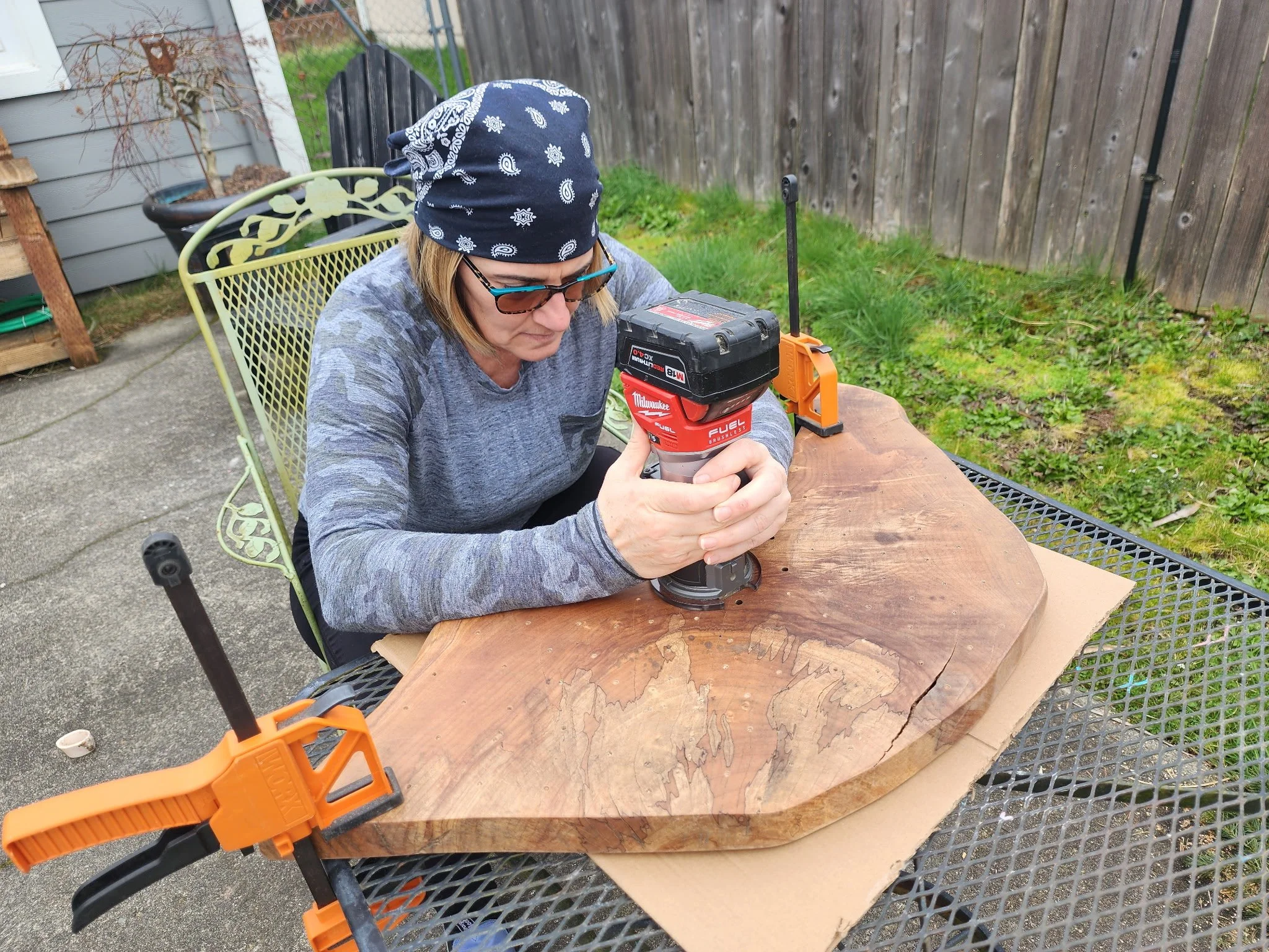 A woman wearing a bandana and sunglasses is working with a rotary tool on a large piece of wood secured with clamps on a metal table outdoors.