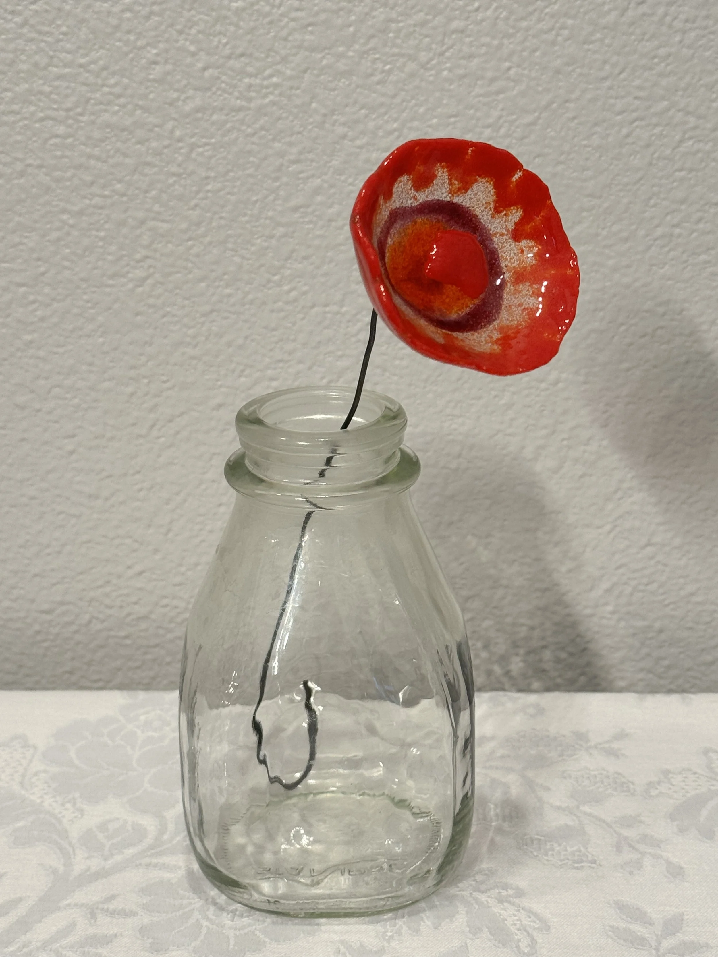 A glass bottle with a flower made from a red poppy with black and white centers, mounted on a thin wire stem.