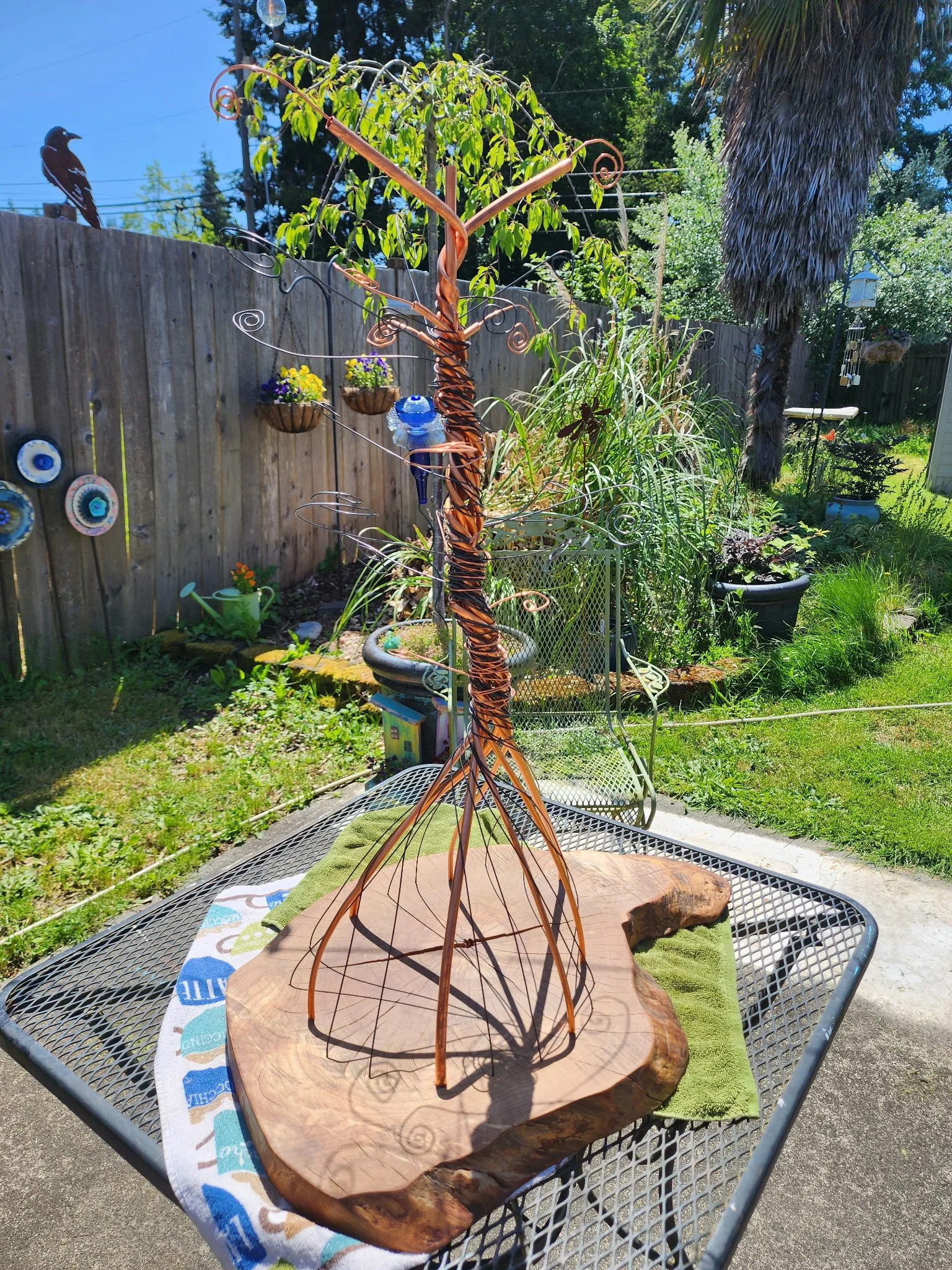 A decorative wire and metal tree sculpture with a wooden base, placed on a outdoor metal table. The sculpture has a twisted metal trunk and wire branches with green leaves. In the background, there are hanging flower pots, a wooden fence, trees, and 