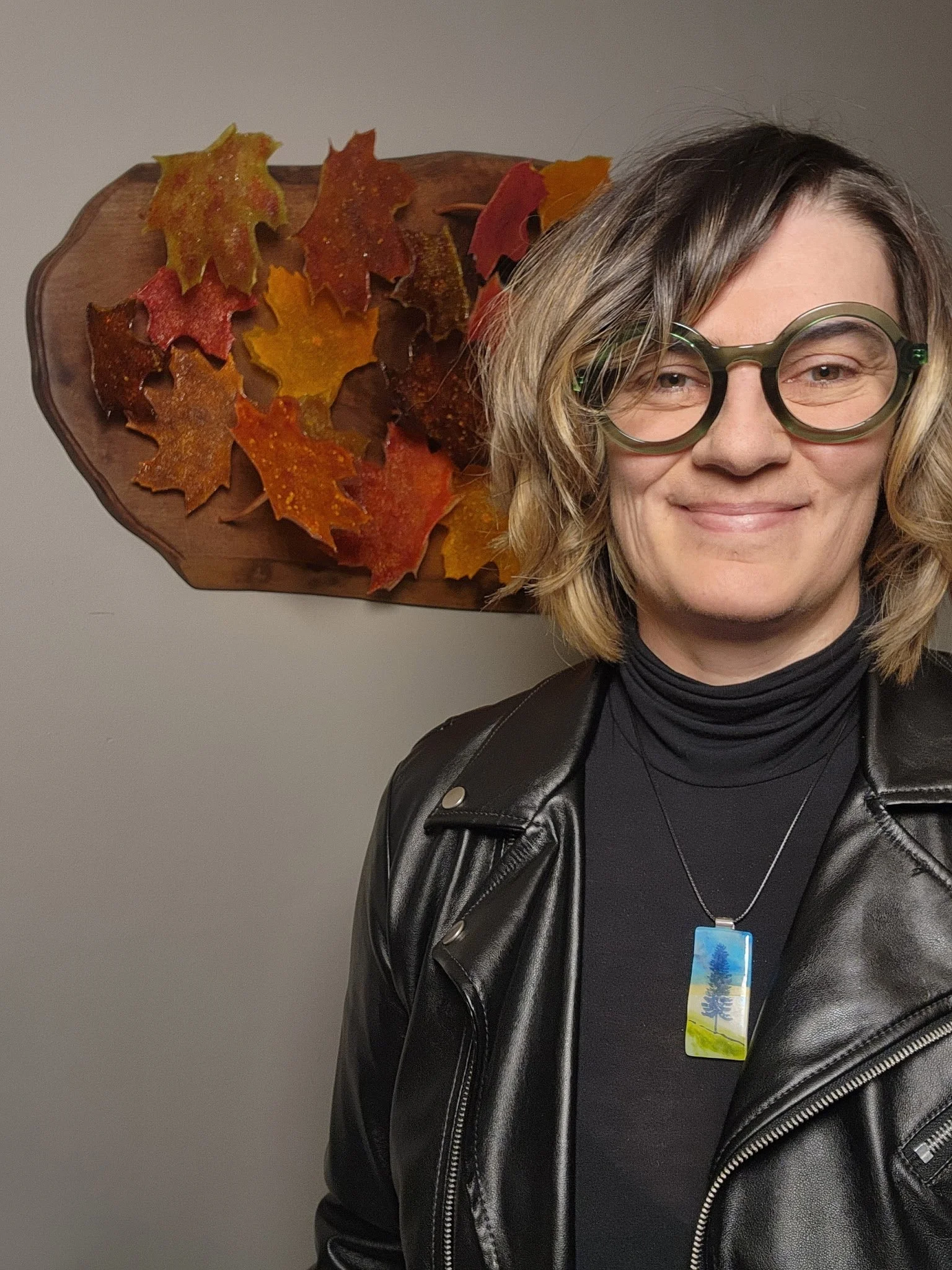 A woman with shoulder-length blond hair wearing large green glasses, a black turtleneck, a leather jacket, and a necklace with a colorful tree pendant, standing in front of a wall with a display of autumn leaves mounted on wood.