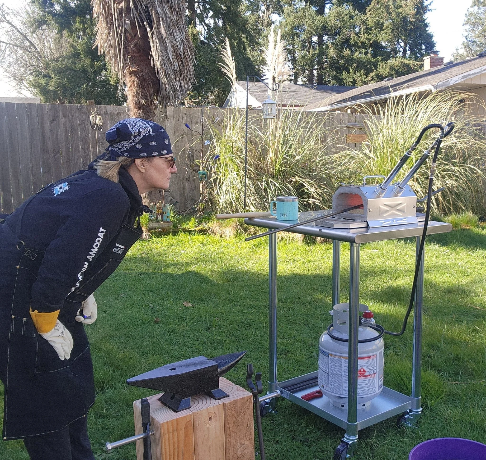 Woman using a blacksmithing forge outdoors, standing with her hands on her hips, looking at a small forge with a steel tool on top, connected to a propane tank, set on a cart beside a table with cups and tools, on a grassy backyard.