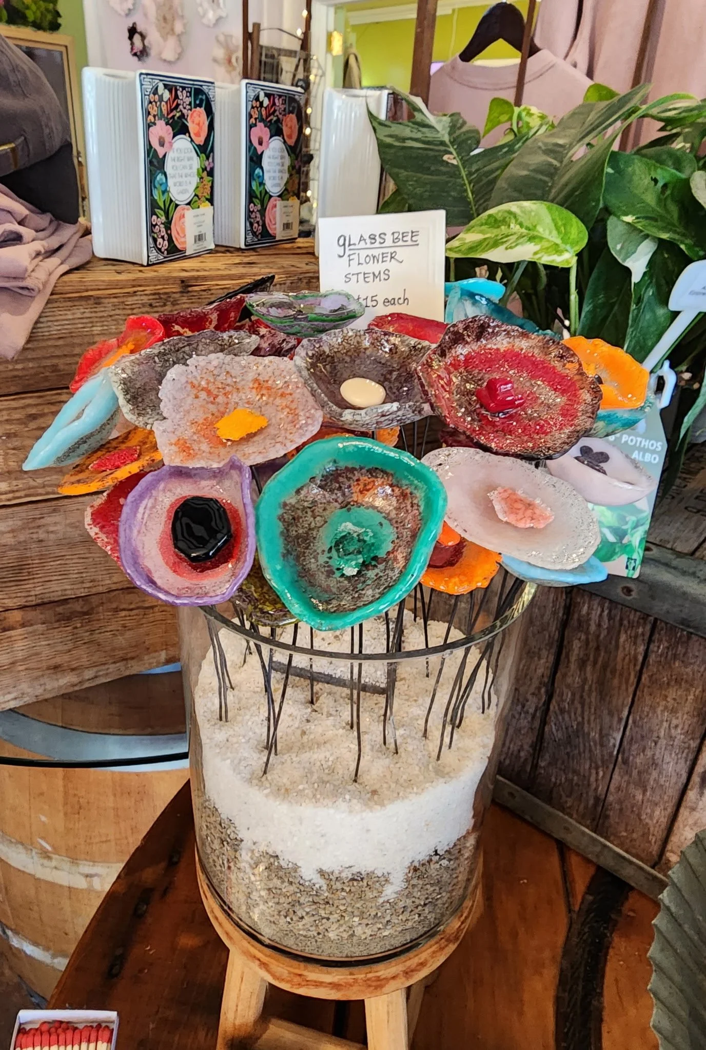 Display of glass bee flower stems for sale, arranged in a wire basket above a sand-filled glass container, in a store setting.