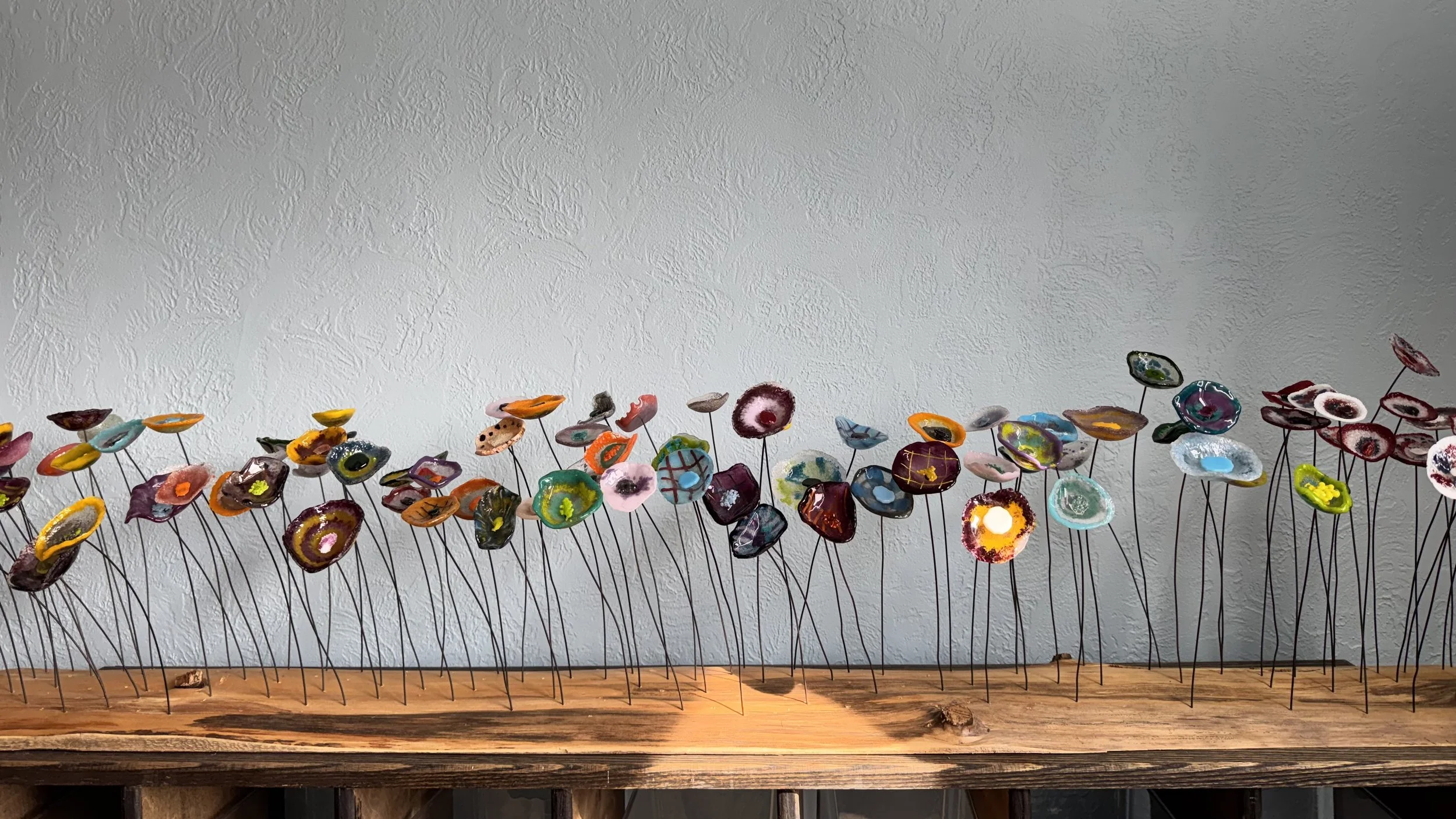 Colorful glass flowers on metal stems arranged on a wooden surface against a textured wall.