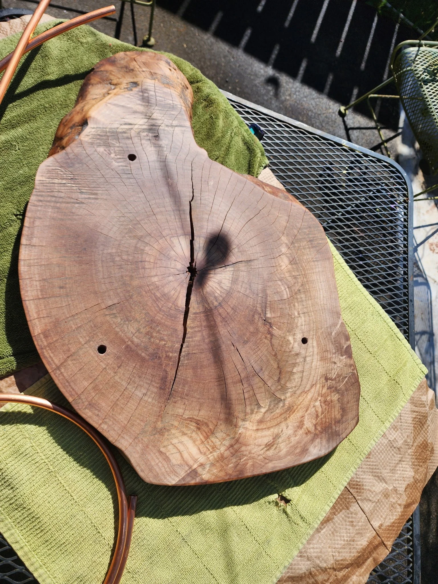 A large round wooden slab with visible growth rings and cracks, placed on a green towel on a metal table outdoors.