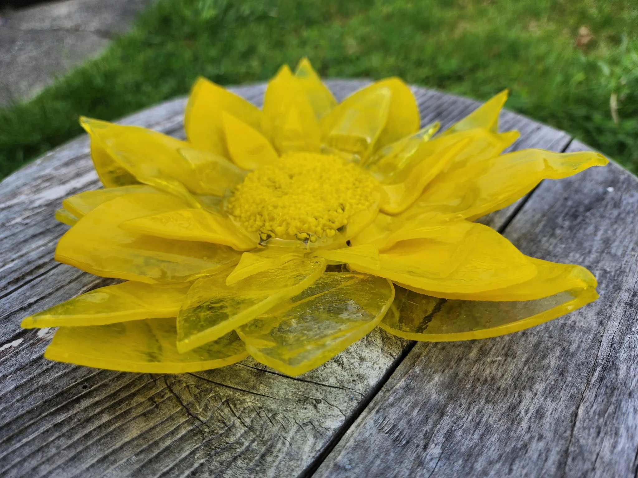 A yellow flower made of glass pieces on a weathered wooden surface with grass in the background.