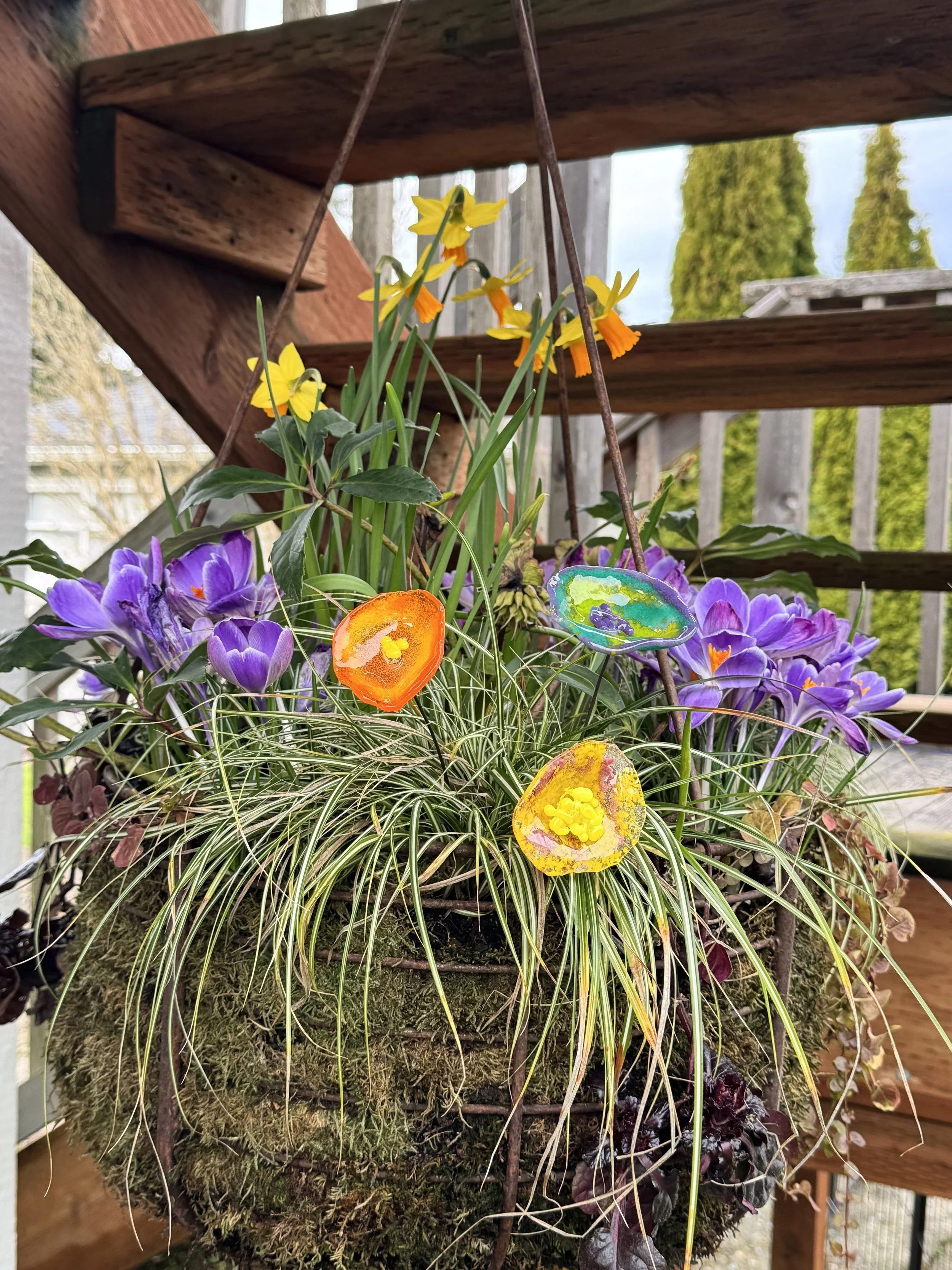 Colorful glass Easter eggs hanging in a flower basket filled with purple crocuses, yellow daffodils, and decorative grass, located on outdoor wooden stairs.