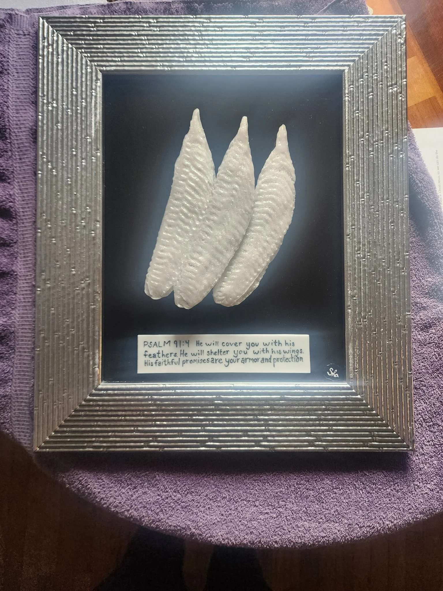 A framed display featuring three white feathers with a note underneath, referencing Psalm 91:4 about God's protection, set on a purple fabric surface.