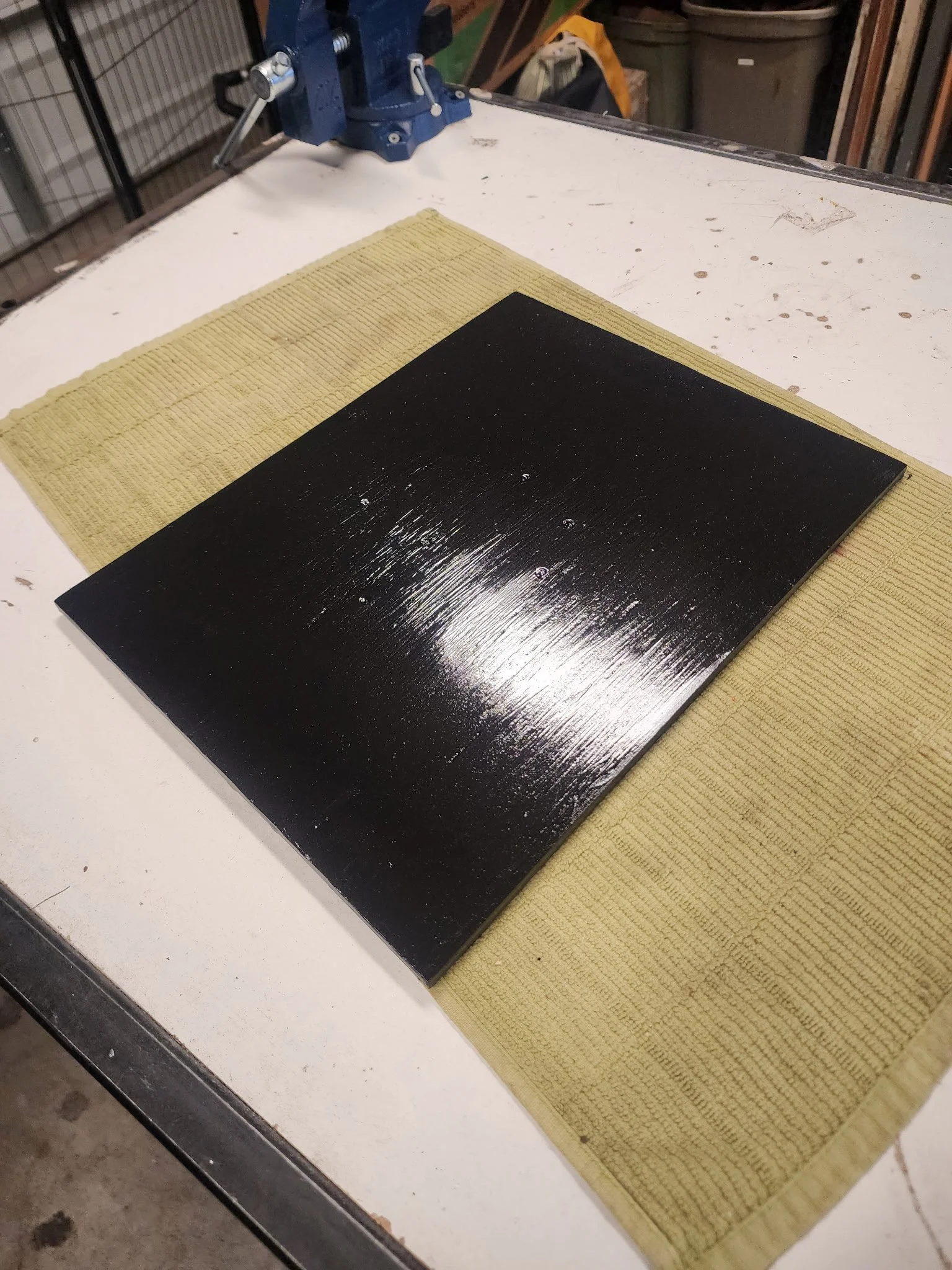 Black painted wooden board with a glossy finish, placed on a beige mat on a white worktable in a workshop.