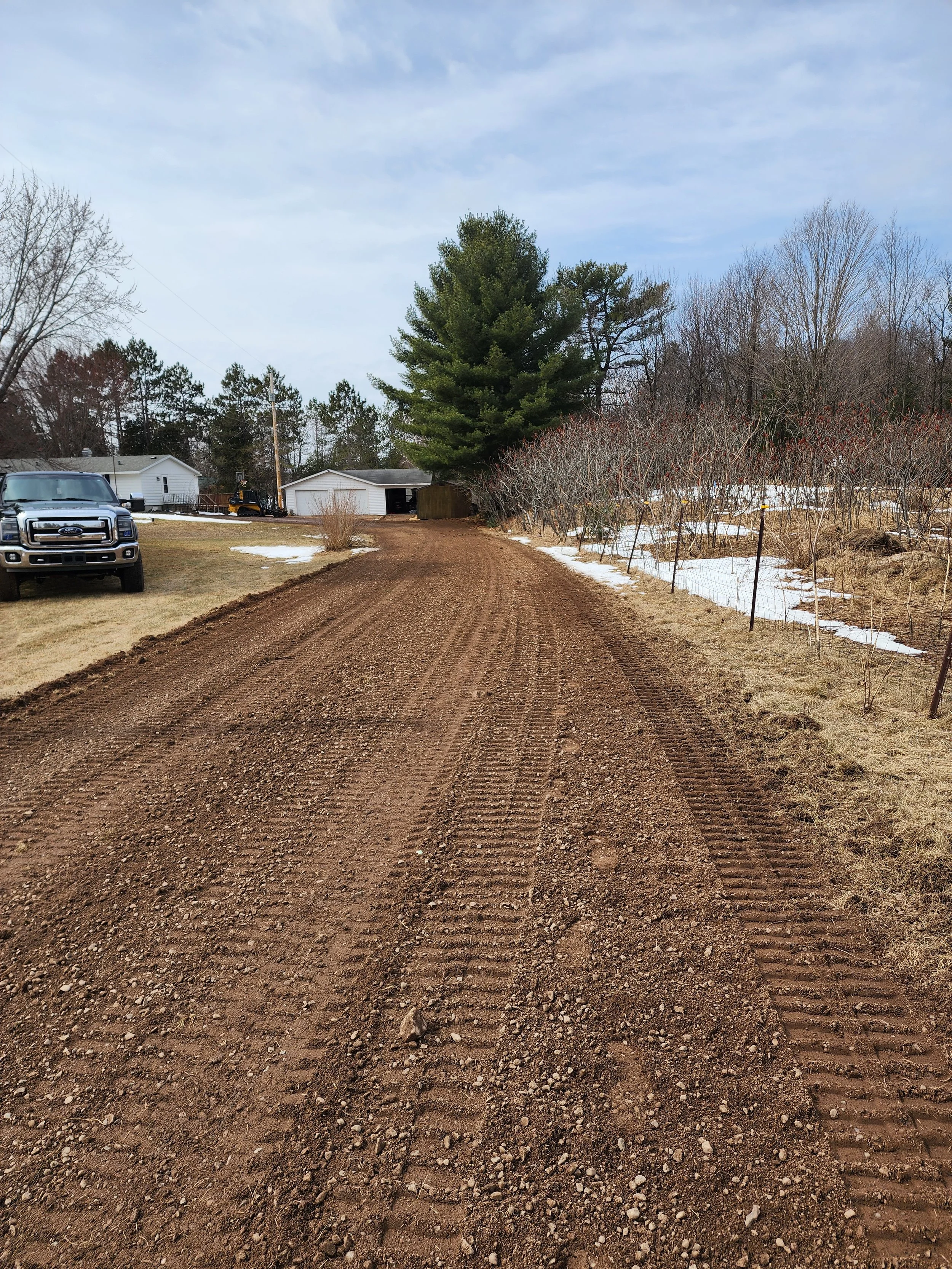 A dirt driveway leading to a garage, with tire tracks, surrounded by patches of snow, grass, trees, and a parked truck.