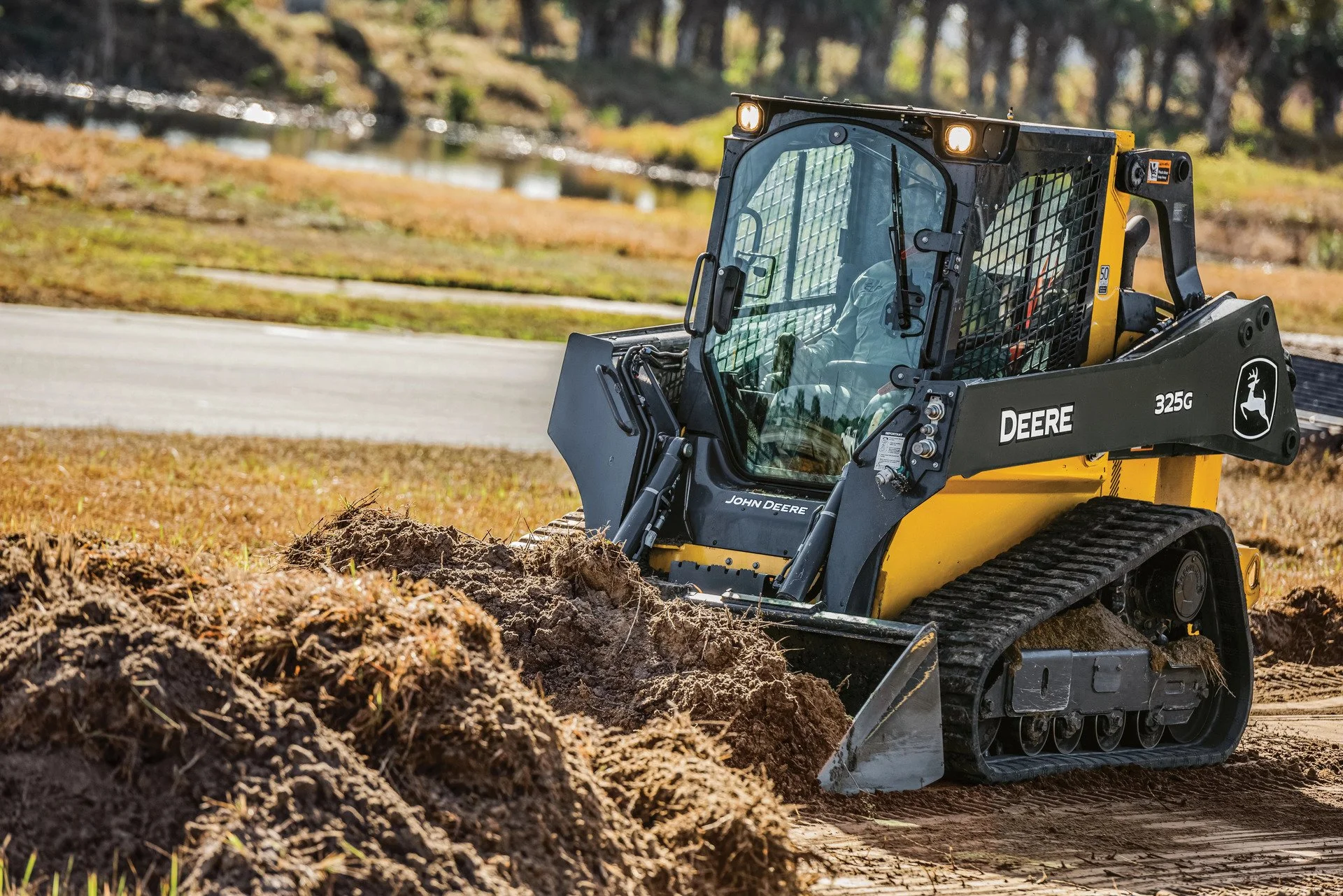 A John Deere compact track loader operating outdoors in a landscaped area, moving soil or dirt near a body of water and surrounded by trees.