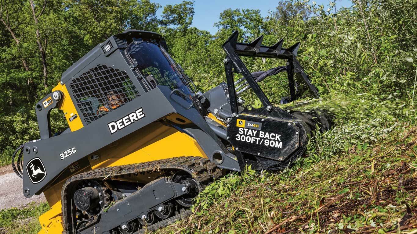 A John Deere 325G compact track loader clearing brush and small trees in a wooded area.