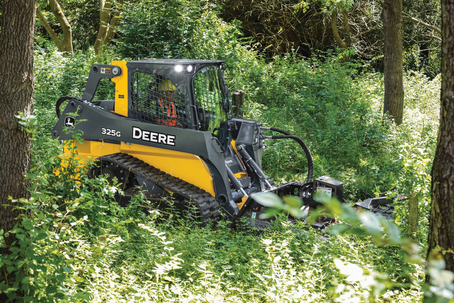 A yellow and black John Deere compact track loader operating in a dense forested area, surrounded by green foliage and trees.