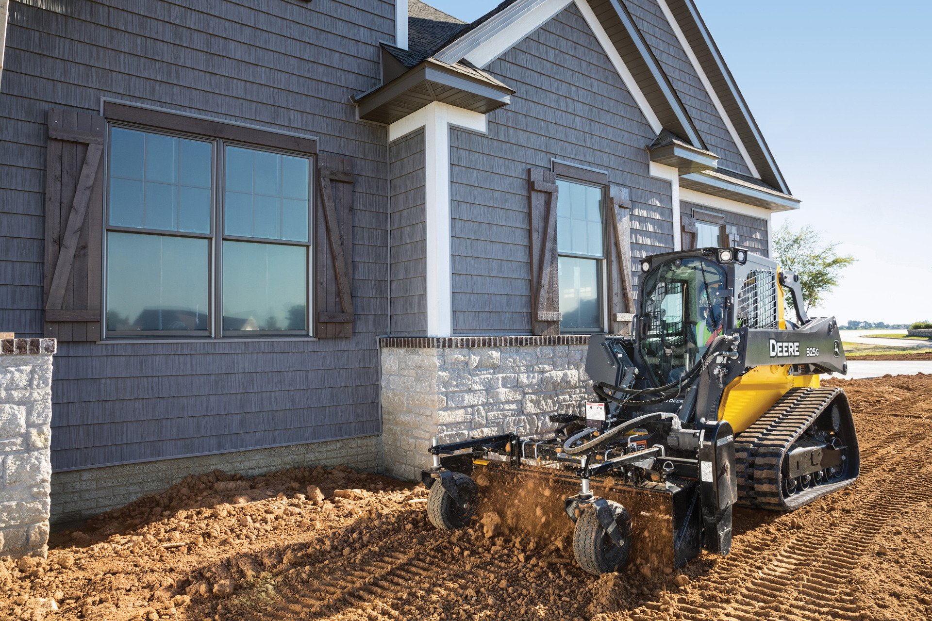 Construction site with a Deere 325G tracked skid steer loader working near a house with gray siding, stone foundation, and large windows.